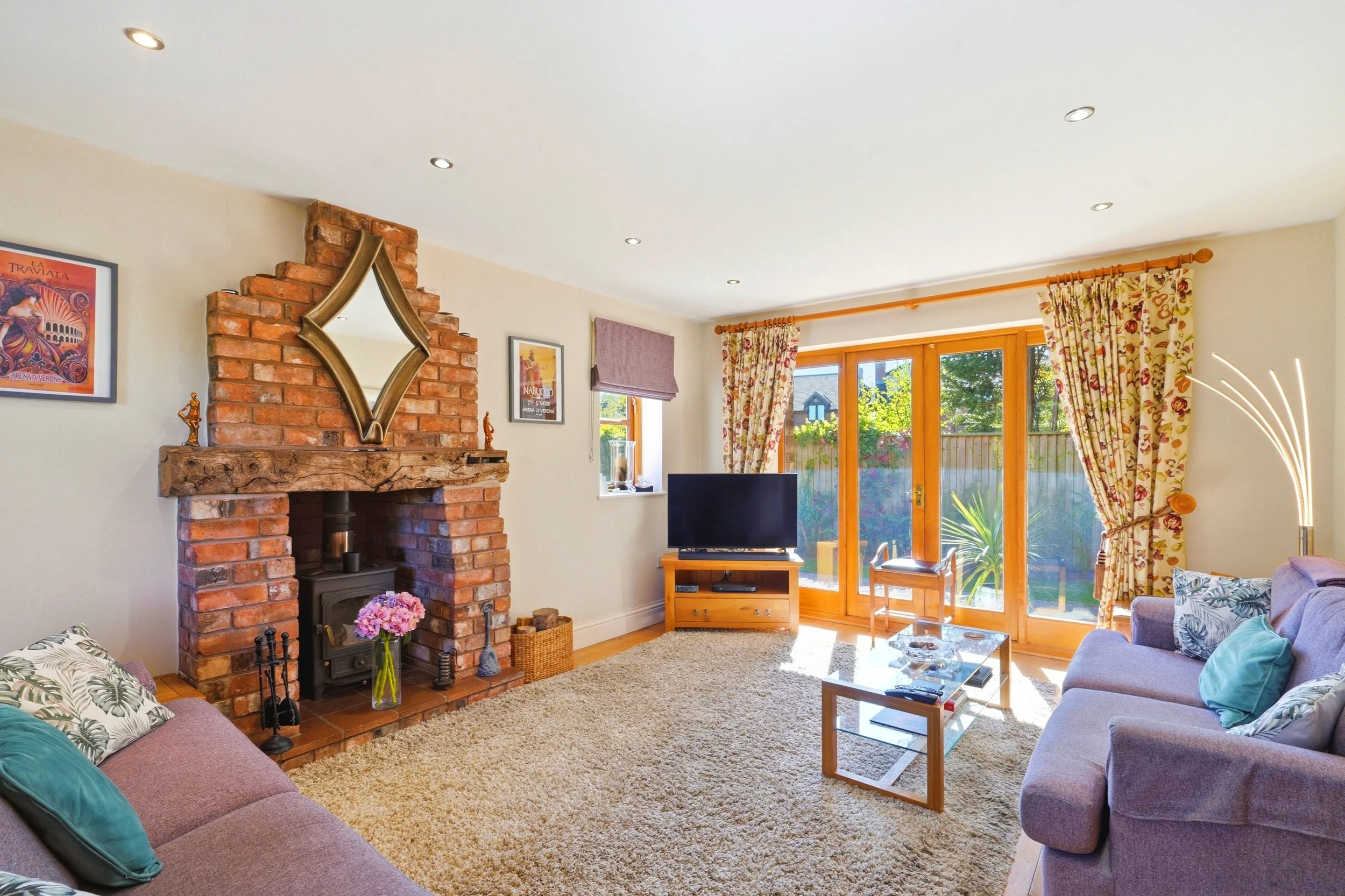 Living room with brick fireplace, wall art, floral curtains, and large glass doors leading outside, with purple and patterned sofas, a TV on a wooden stand, and a beige shag rug.