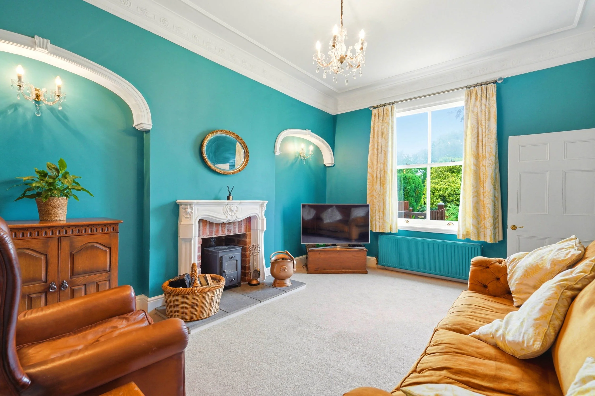 A living room with teal walls, a white painted fireplace, a flat-screen TV, a wooden cabinet, a leather armchair, a yellow sofa with pillows, a large window with yellow curtains, a chandelier, wall sconces, a potted plant, and various decorative item