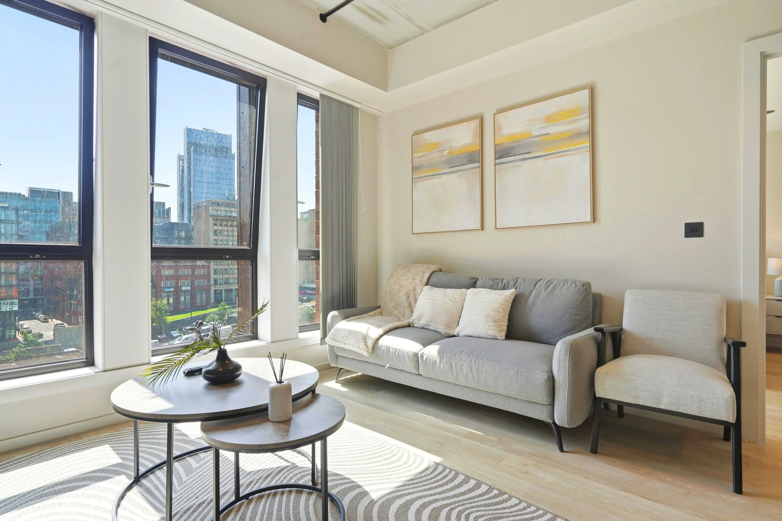 Modern living room with large windows showing city buildings, a grey sofa with cushions, a round coffee table with decorative items, a striped rug, and wall art with abstract yellow, grey, and white patterns.