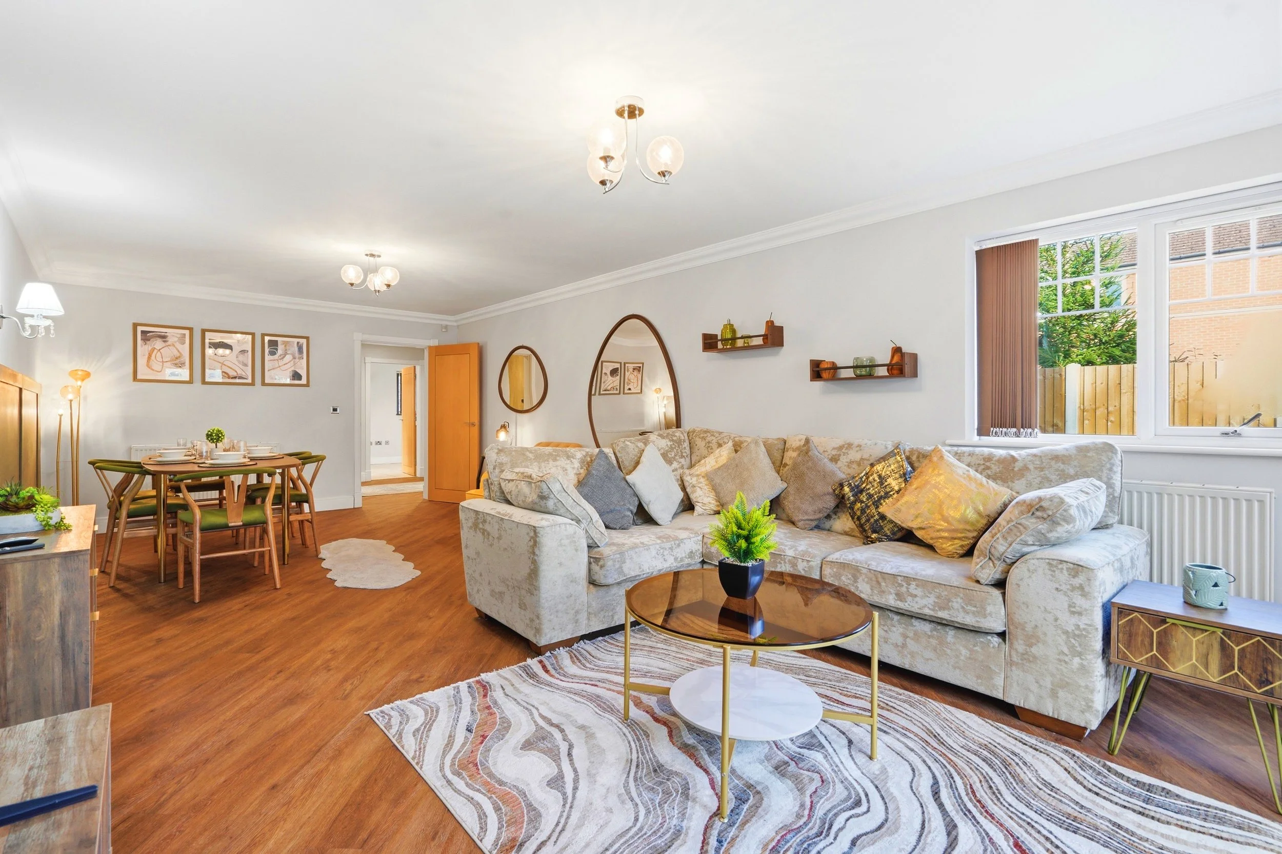 Living room with cream-colored sofa, round glass coffee table, wooden dining table with green chairs, decorative wall shelves, large window, and wooden flooring.
