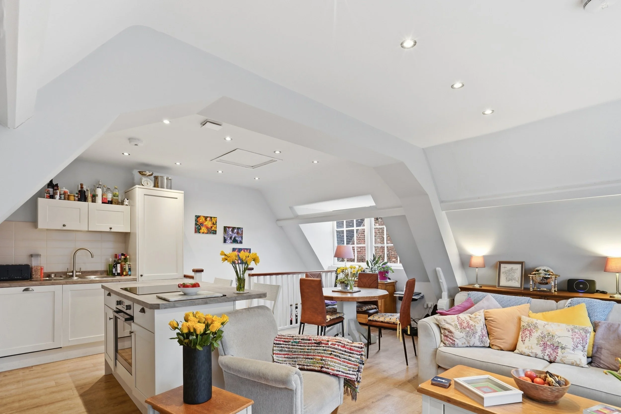 A spacious, open-concept living area with a high sloped ceiling, white walls, and large windows. The space is decorated with colorful artwork, floral arrangements, and cozy furniture including a sofa with pillows, chairs, and a dining table. The kitc