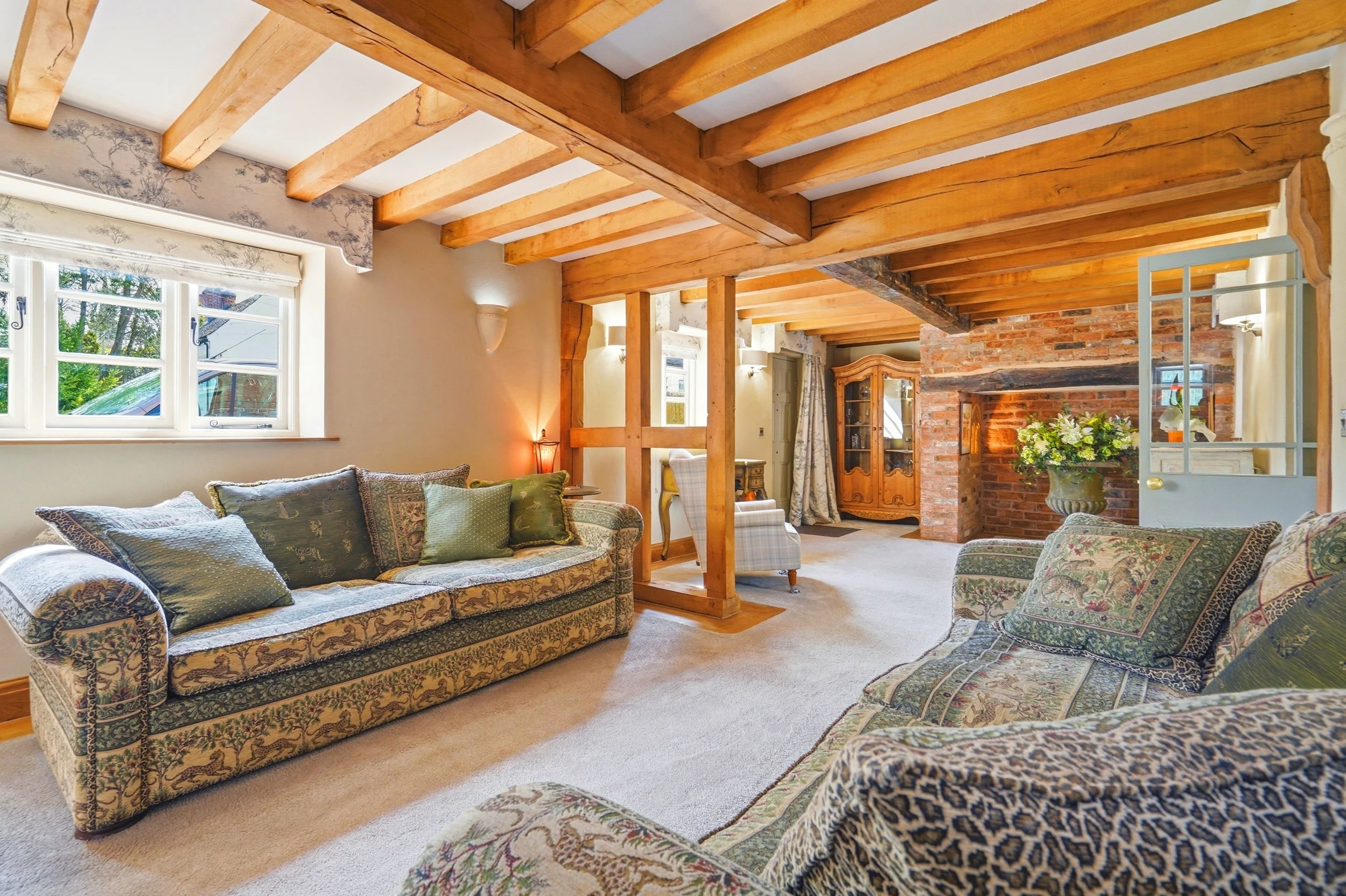 Living room with floral-patterned sofas, wooden beams on the ceiling, a brick fireplace with a floral arrangement, multiple windows allowing natural light, and vintage furniture.