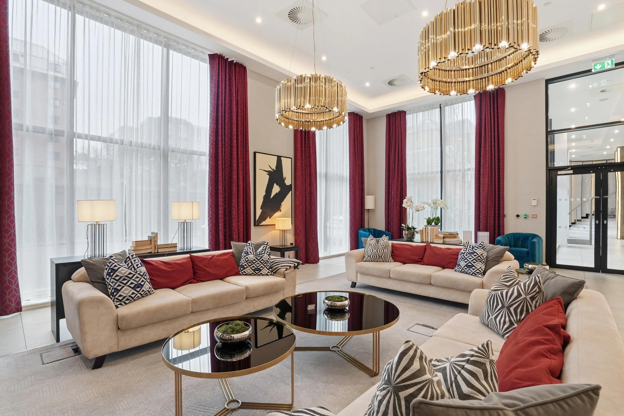 A modern hotel lobby with large windows, red drapes, beige sofas with patterned cushions, black coffee tables, and elegant gold chandeliers.