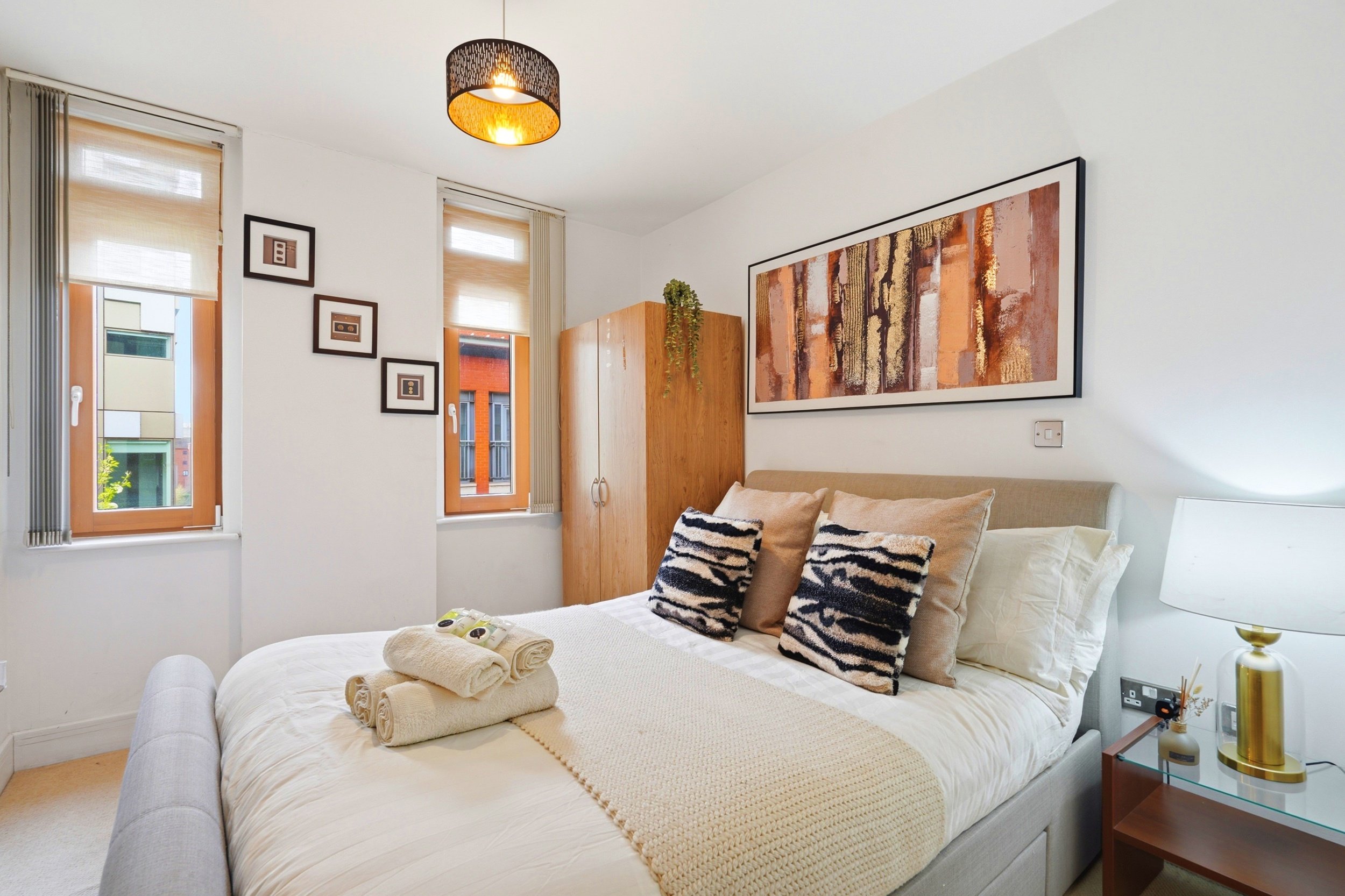 A cozy bedroom with a bed decorated with throw pillows, neatly folded towels, and a beige blanket. Two windows with beige Roman shades let in natural light. Artwork hangs above the bed, and a wooden wardrobe stands in the corner. A modern pendant lig