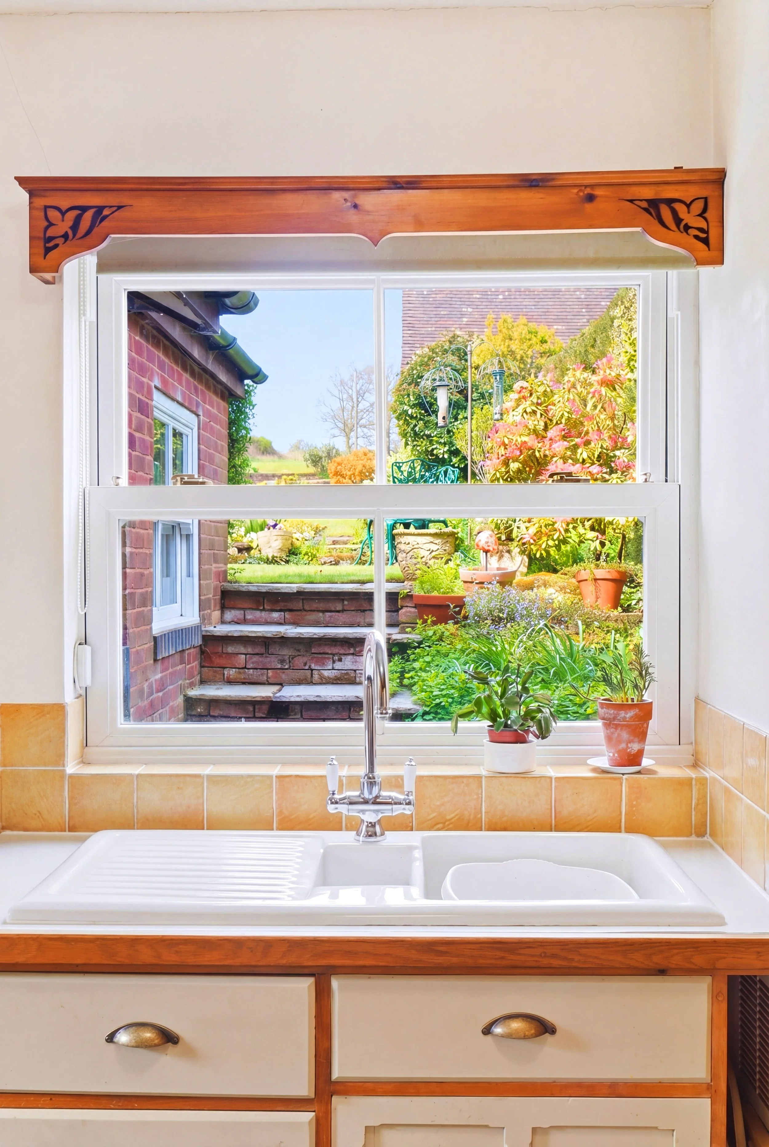 Kitchen sink with a window view of a garden with potted plants, flowers, and trees, and a brick patio outside.