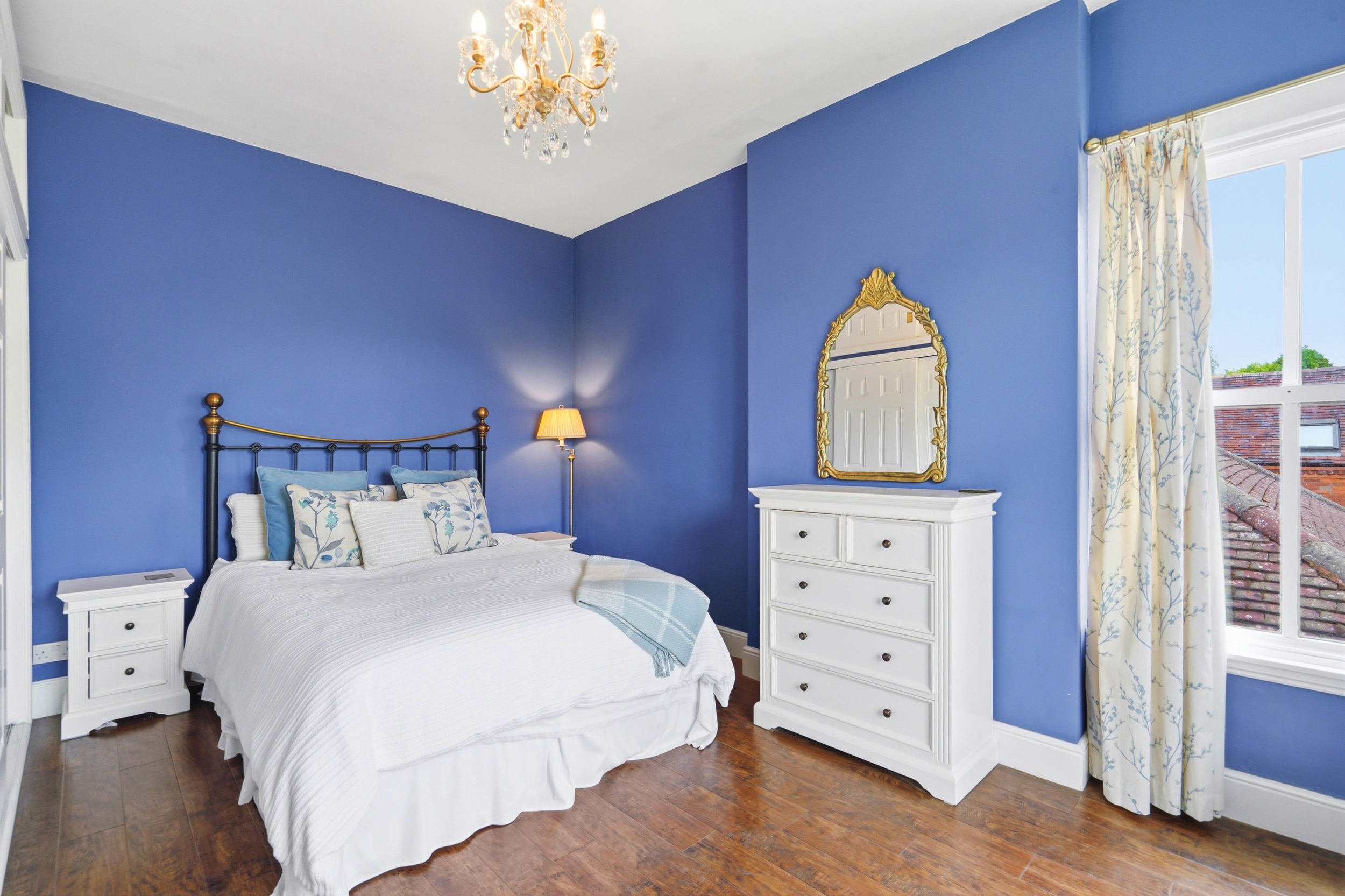 A bedroom with a white bed, blue walls, and a window with floral curtains. There is a gold-framed mirror above a white dresser, a small white nightstand with a drawer, and a chandelier hanging from the ceiling.