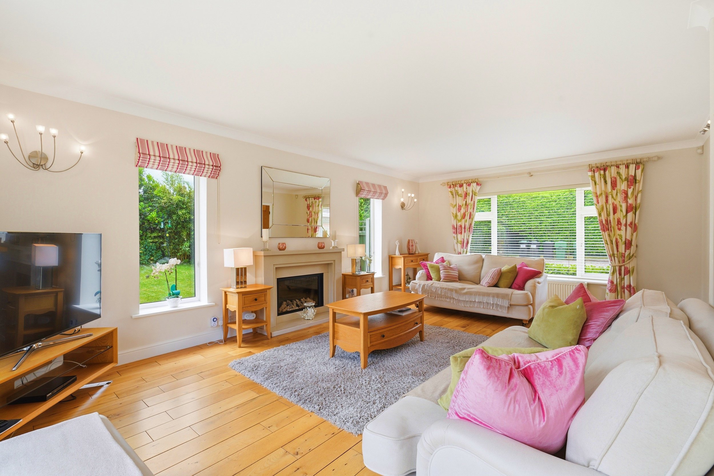 Living room with a white sofa, green and pink cushions, wooden furniture, a gray rug, big windows with pink floral curtains, a fireplace, and a flat-screen TV.