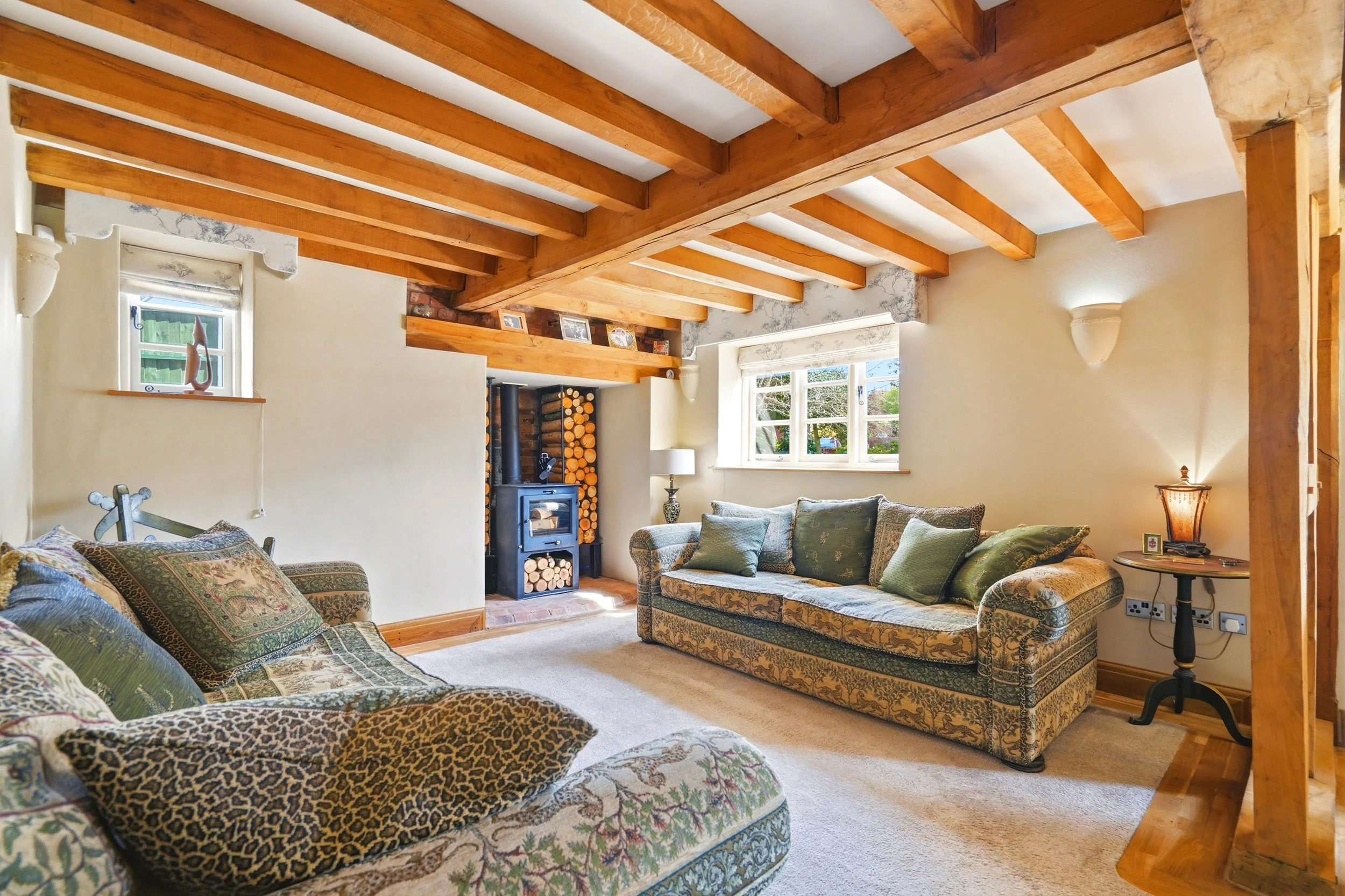 Cozy living room with wooden ceiling beams, floral-patterned furniture, a brick fireplace with stacked logs, a window with white curtains, and warm lighting.