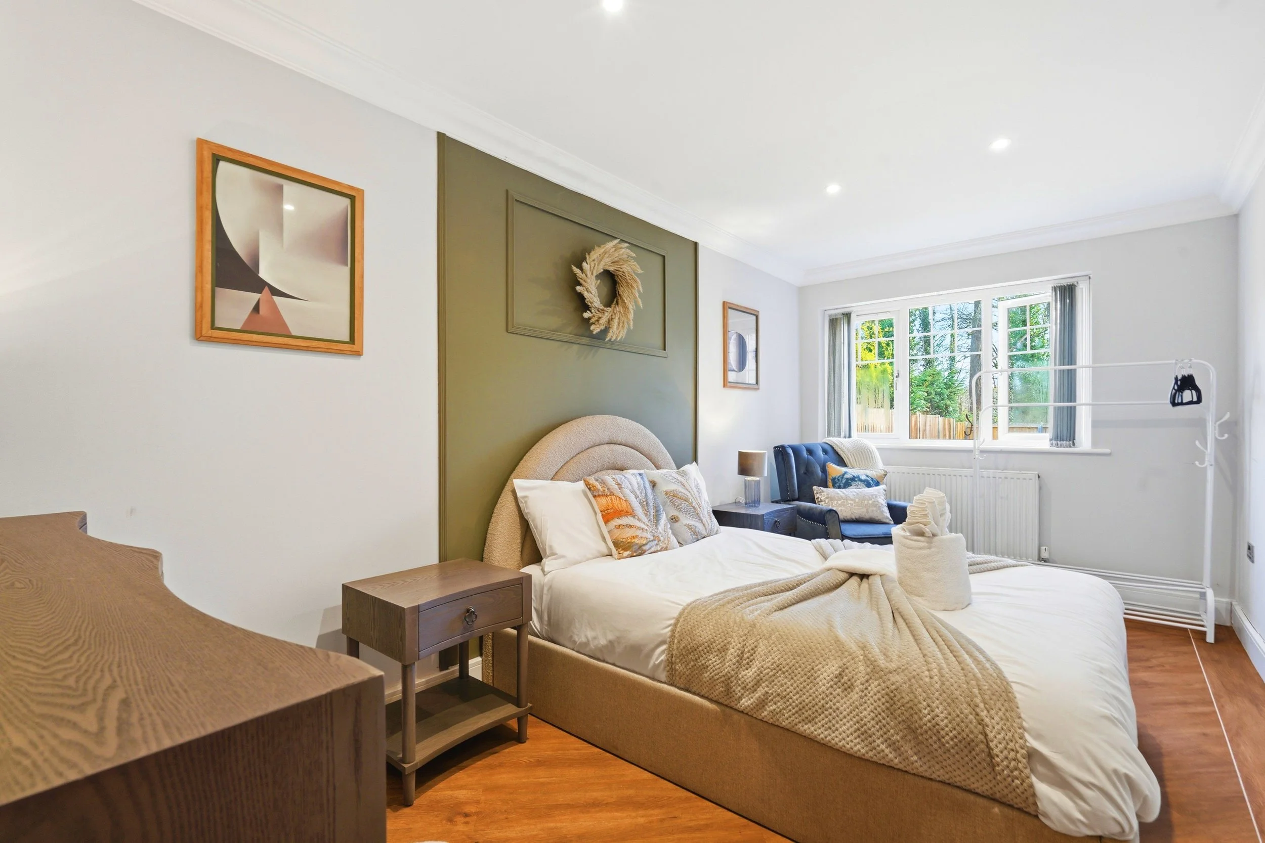 A bright bedroom with a large window, a bed with pillows and a beige duvet, a nightstand with a lamp, artwork on the walls, and a green accent wall with a decorative wreath.