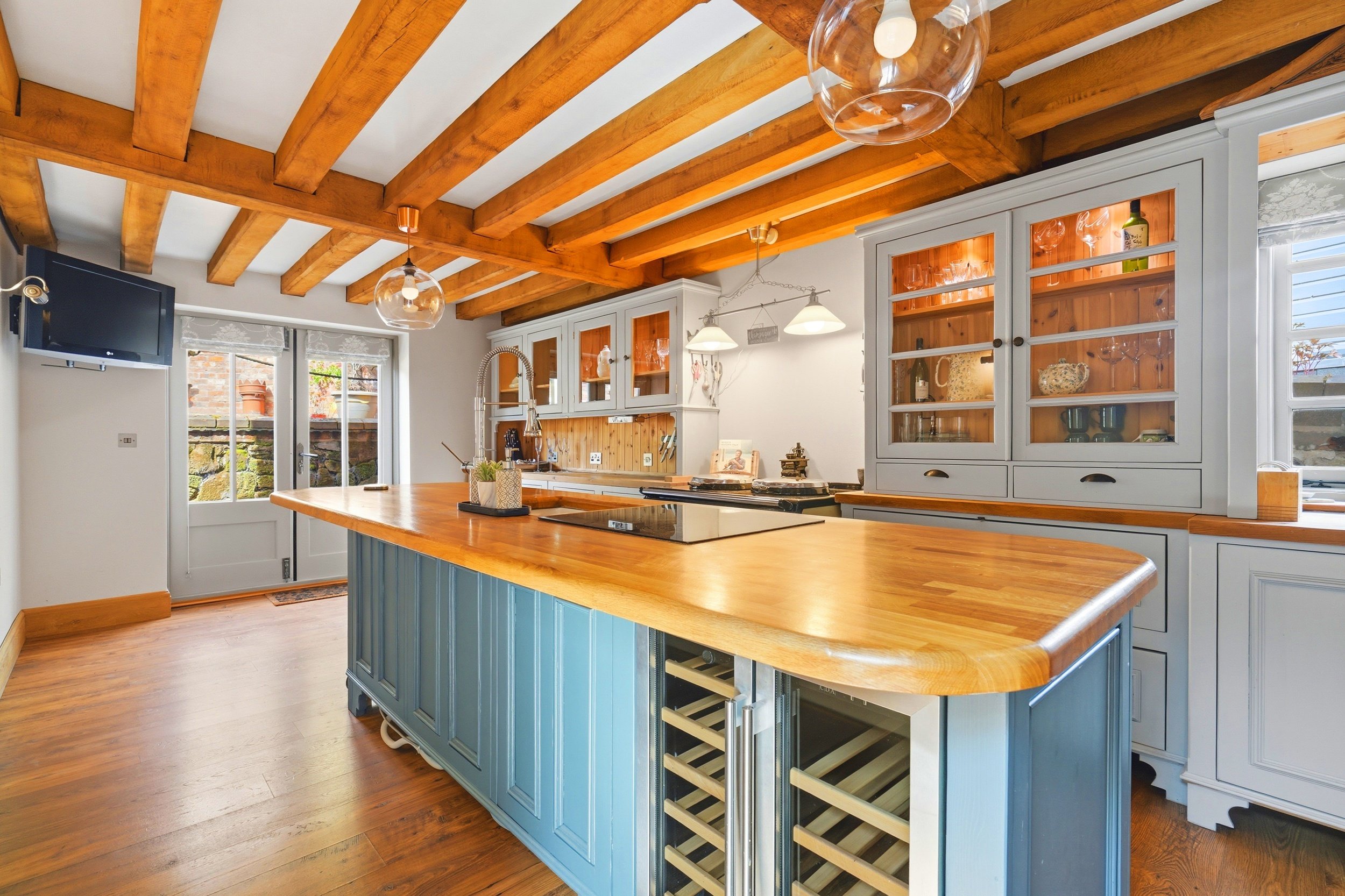 A kitchen with a wooden ceiling and exposed beams, light blue cabinets, and a central island with a wooden countertop. There is a wine fridge built into the island, a sink with a sprayer faucet, and glass-front cabinets showing dishes inside. A door 