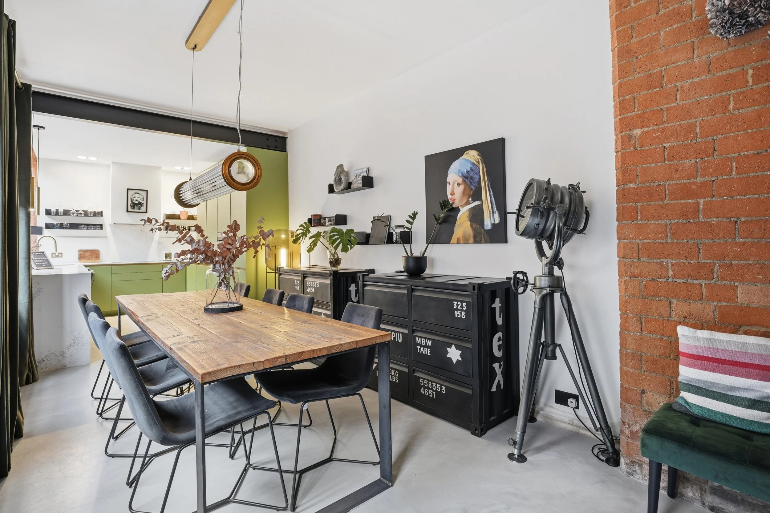 A modern dining room with a large wooden table surrounded by black chairs, a brick wall partially on the right, a black cabinet with decorative items and plants, a large portrait of a woman in a headscarf on the wall, and a vintage spotlight on a tri