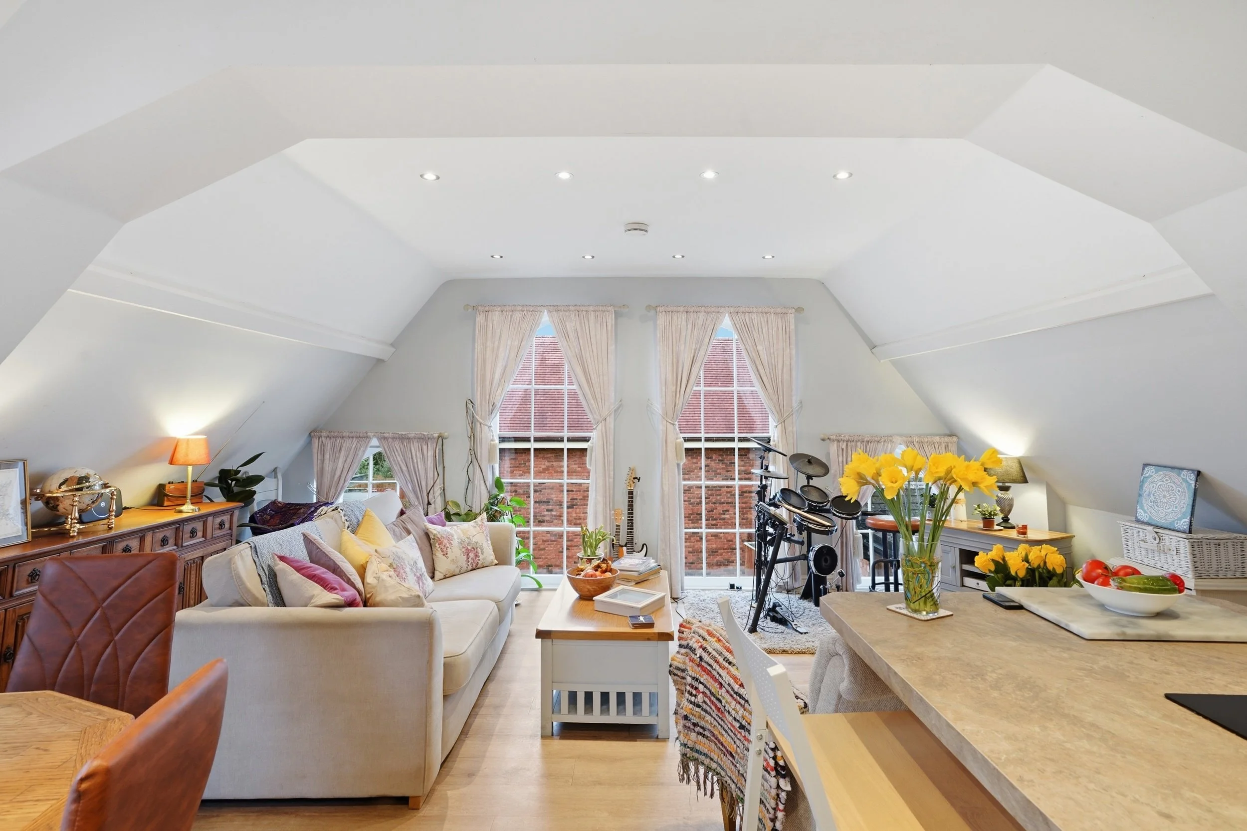 A bright living room with white walls and sloped ceiling, large windows with curtains, a sofa with multiple pillows, a wooden sideboard, and plants. There is a table with a flower vase, and musical instruments near the windows. The room is cozy and w