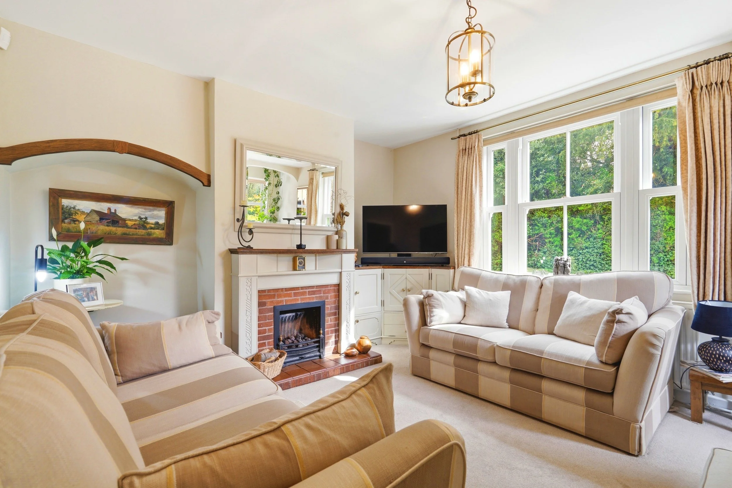 Living room with beige sofas, a fireplace, a flat-screen TV, large windows with curtains, and decorative items.