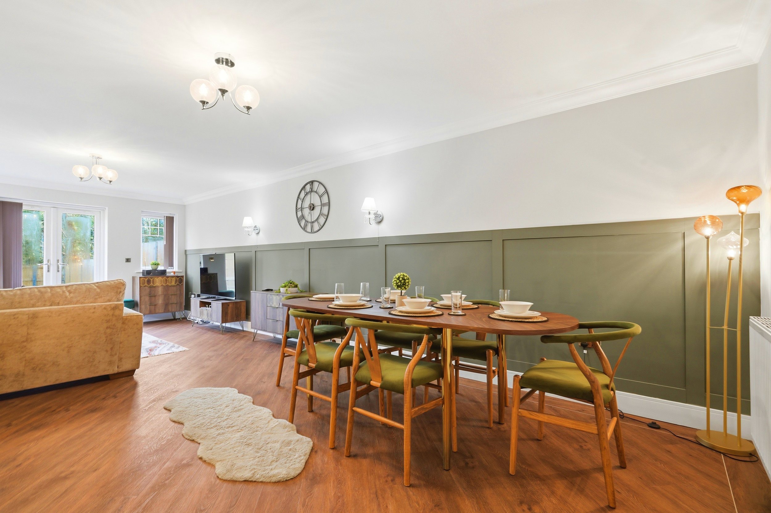 Living and dining area with wooden dining table set with bowls and glasses, green cushioned chairs, beige sofa, wall clock, wall-mounted lamps, and pendant lights