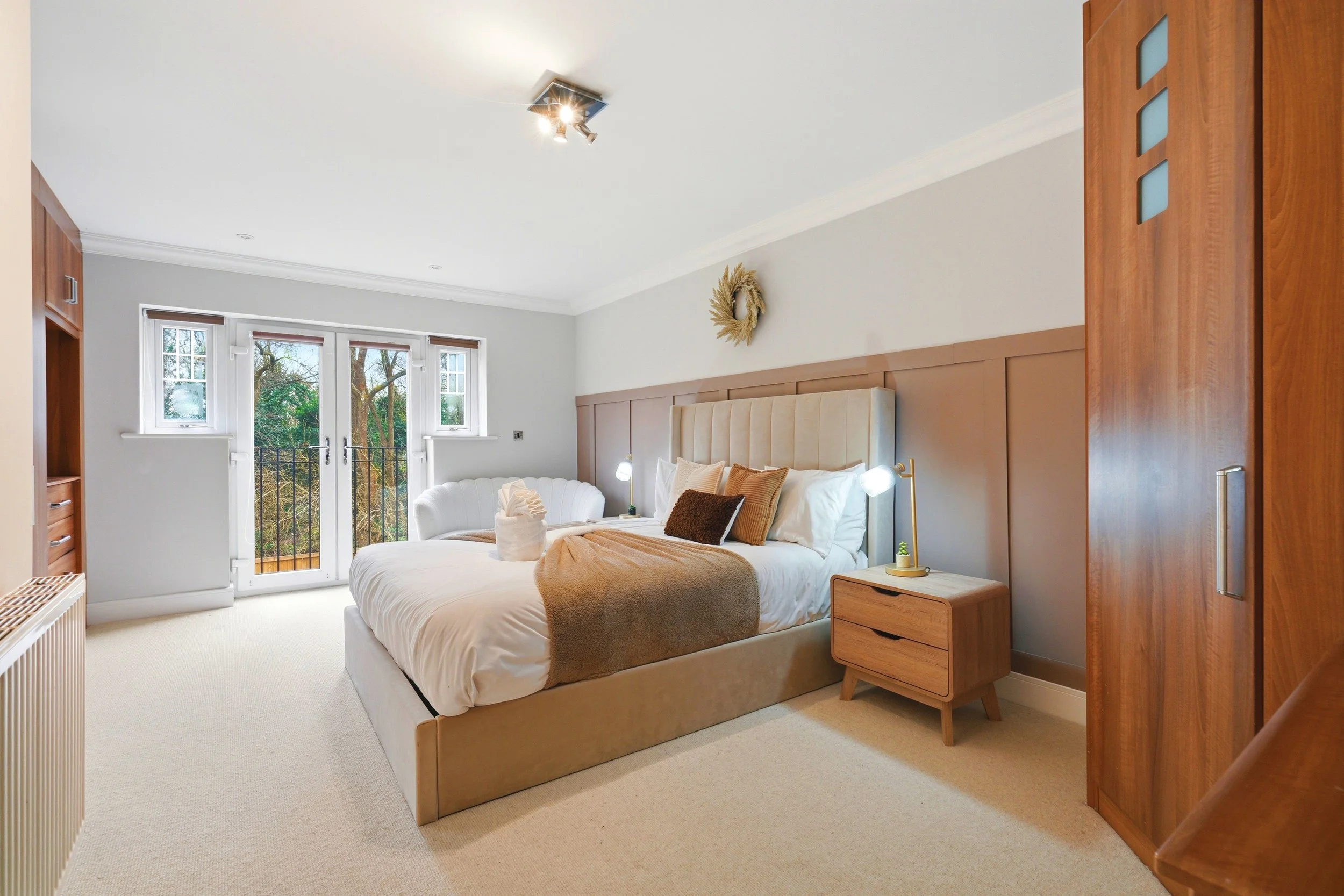 A cozy bedroom with a large bed dressed in white linens, tan and brown pillows, and a brown throw blanket. There is a wooden nightstand with a small potted plant and lamp. The room has large windows with a view of trees outside, and a door leading to