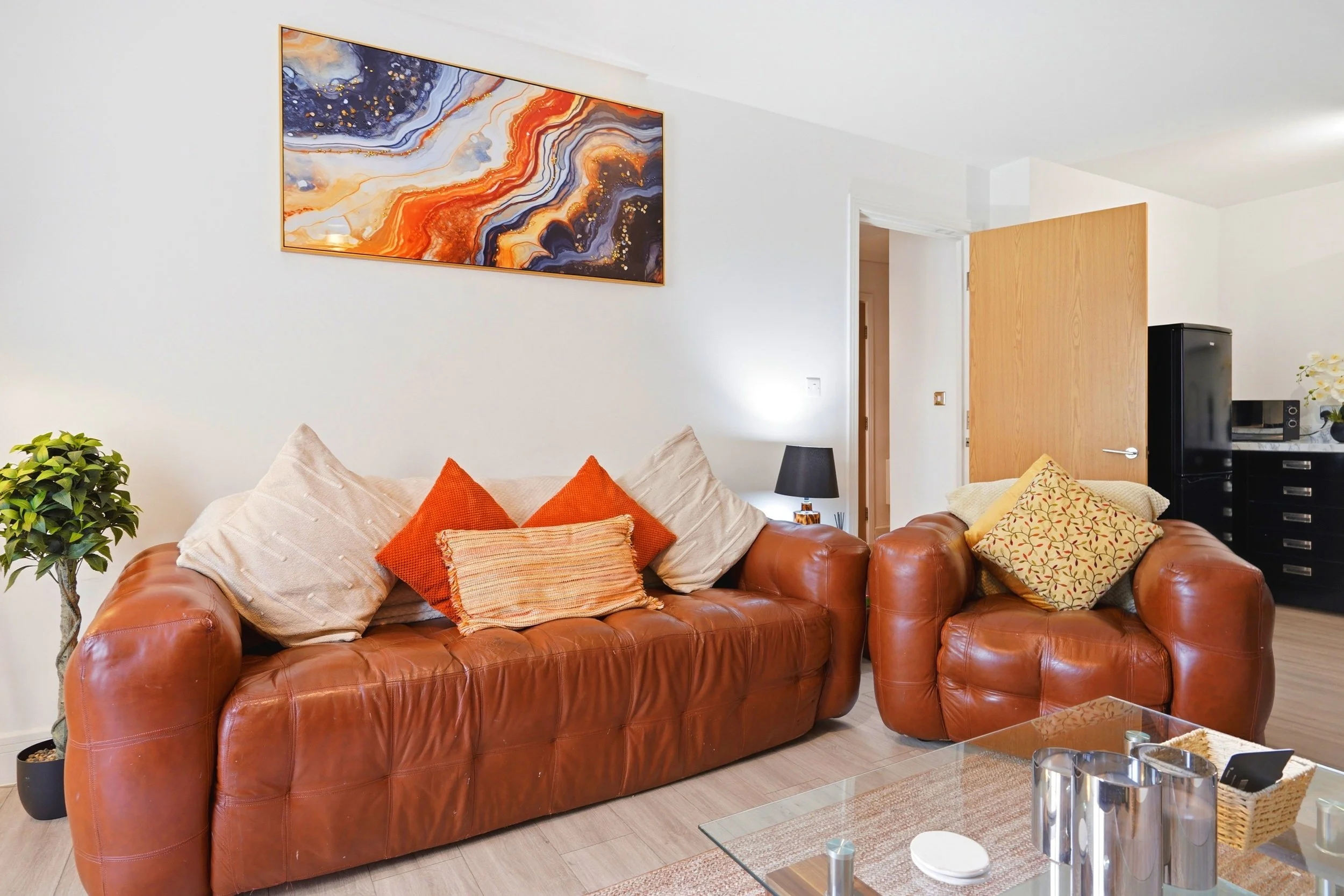 A living room with a leather sofa decorated with beige and orange cushions, a glass coffee table, a potted plant, and a colorful abstract painting on the wall.