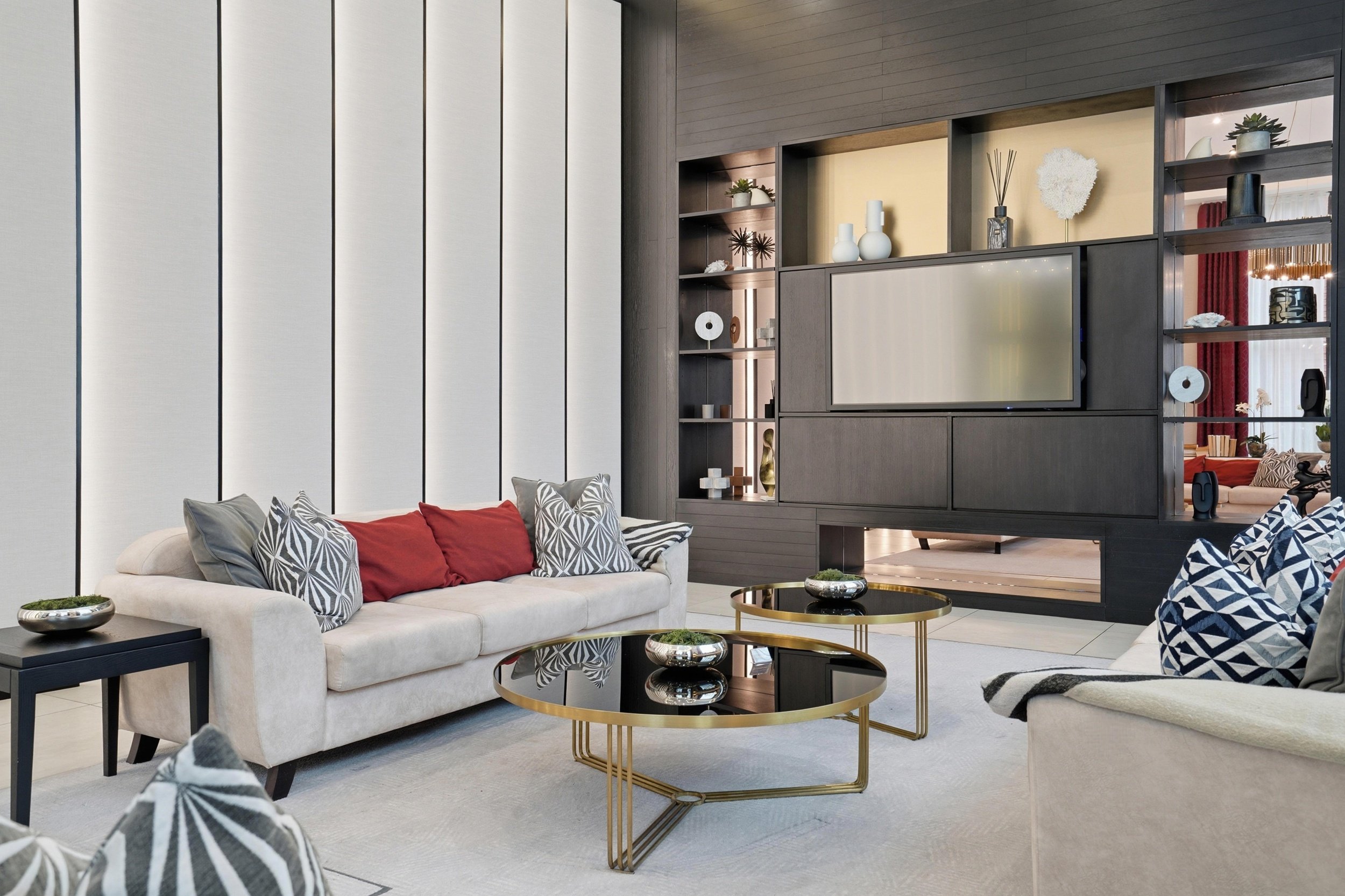 Modern living room with white sofa, patterned pillows, black side table, gold and black coffee tables, gray built-in shelves with decorative items, a large flat-screen TV, and a gray and beige color scheme.