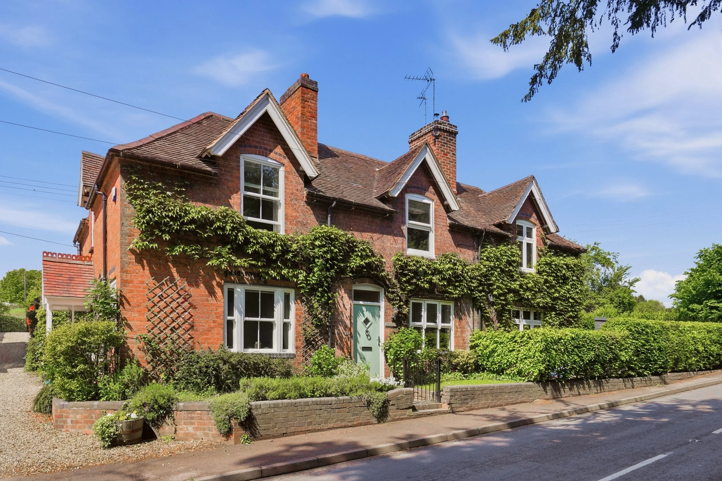 A brick house with multiple gabled roofs and two brick chimneys, surrounded by greenery and a low brick wall, under a clear blue sky.