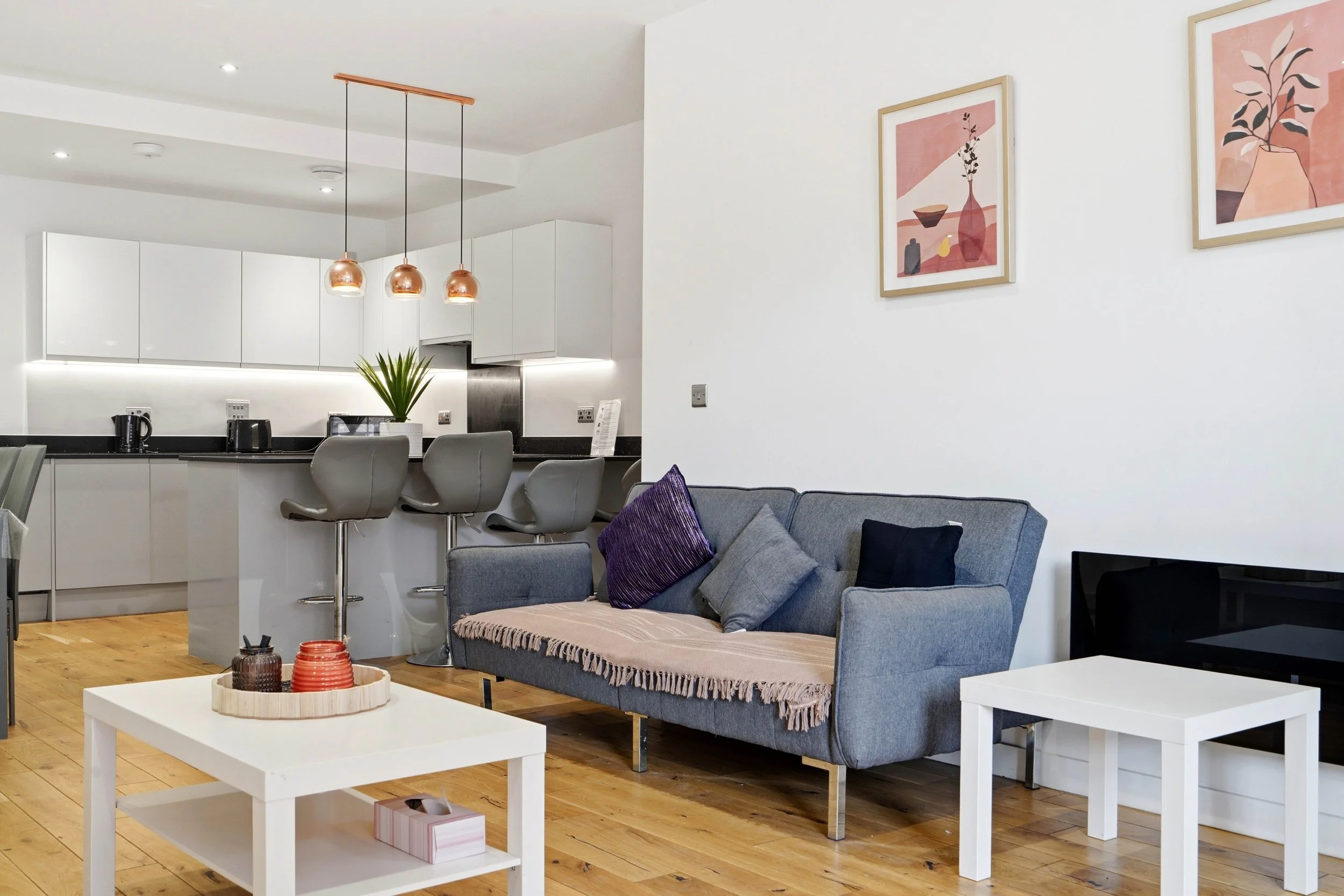 A modern living room and kitchen area with white cabinetry, copper pendant lights, a gray sofa with purple and black throw pillows, a white coffee table with decorative objects, art on the wall, and hardwood flooring.
