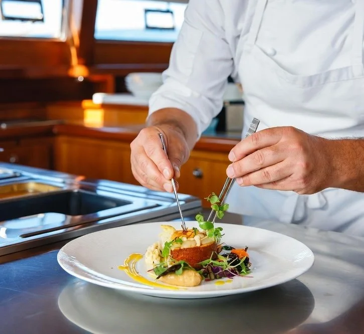 Chef garnishing a plated dish with herbs and sauces in a professional kitchen.