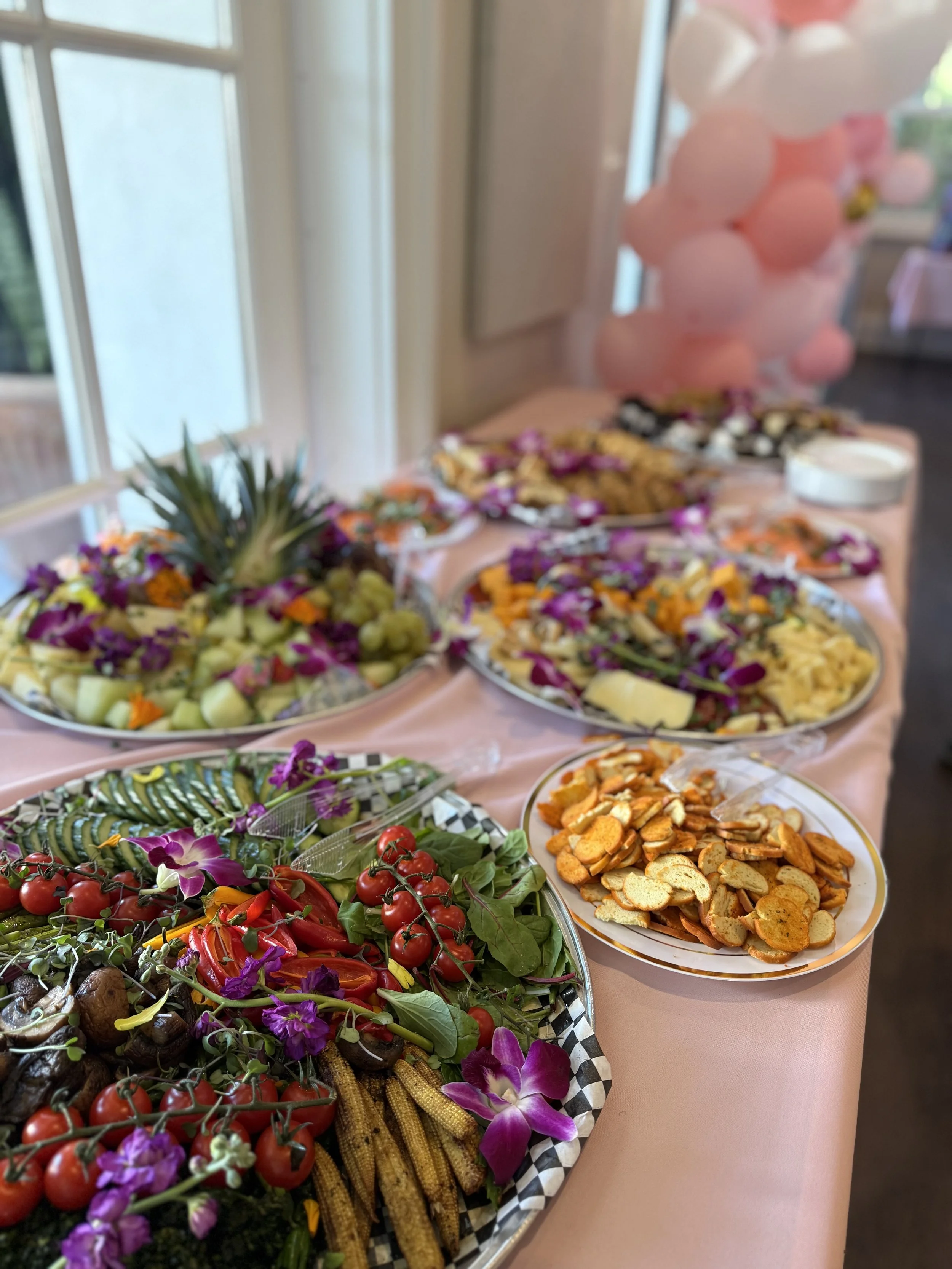 A table with various foods and snacks, including cherry tomatoes, flowers, and crackers, with pink balloons in the background.