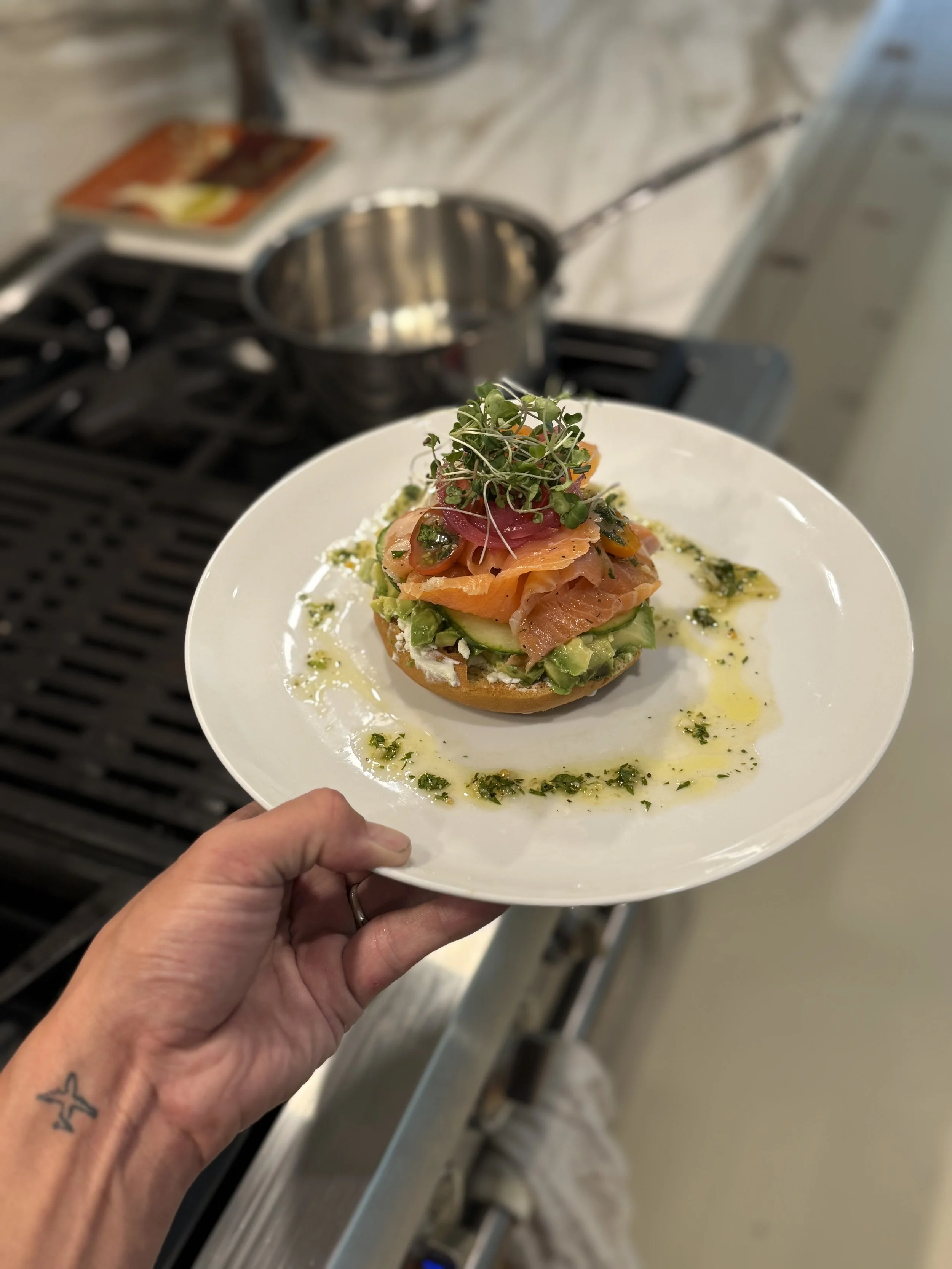 Hand holding a white plate with an open-faced sandwich topped with salmon, microgreens, and green sauce. In the background, a stove with a pot and a book on the counter, slightly out of focus.