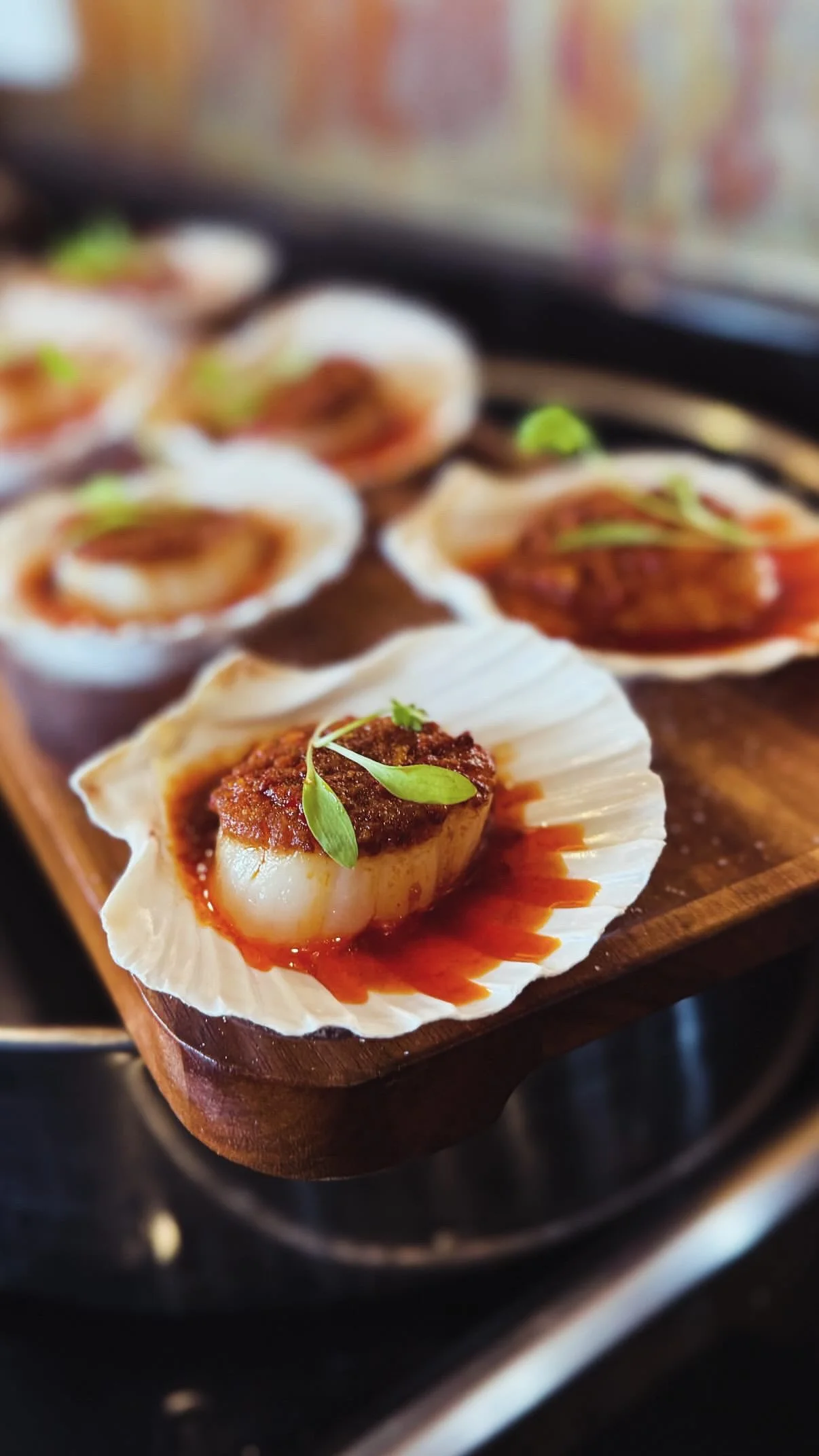 Close-up of a baked scallop topped with sauce and herbs, served in a shell on a wooden serving board.