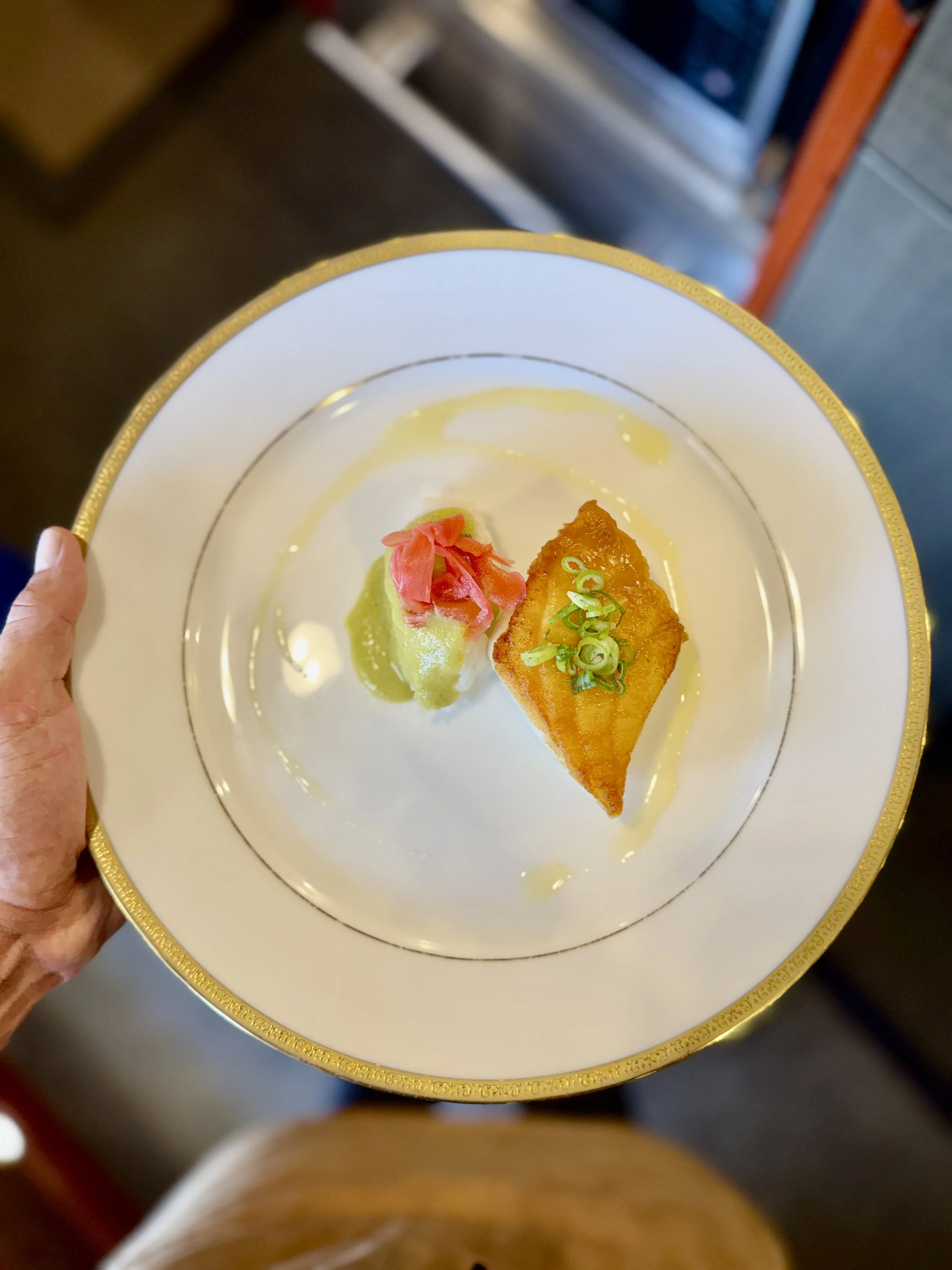 Hand holding a decorative white plate with gold trim, featuring a piece of fried fish topped with green onions, served with a side of green sauce and pickled pink ginger.