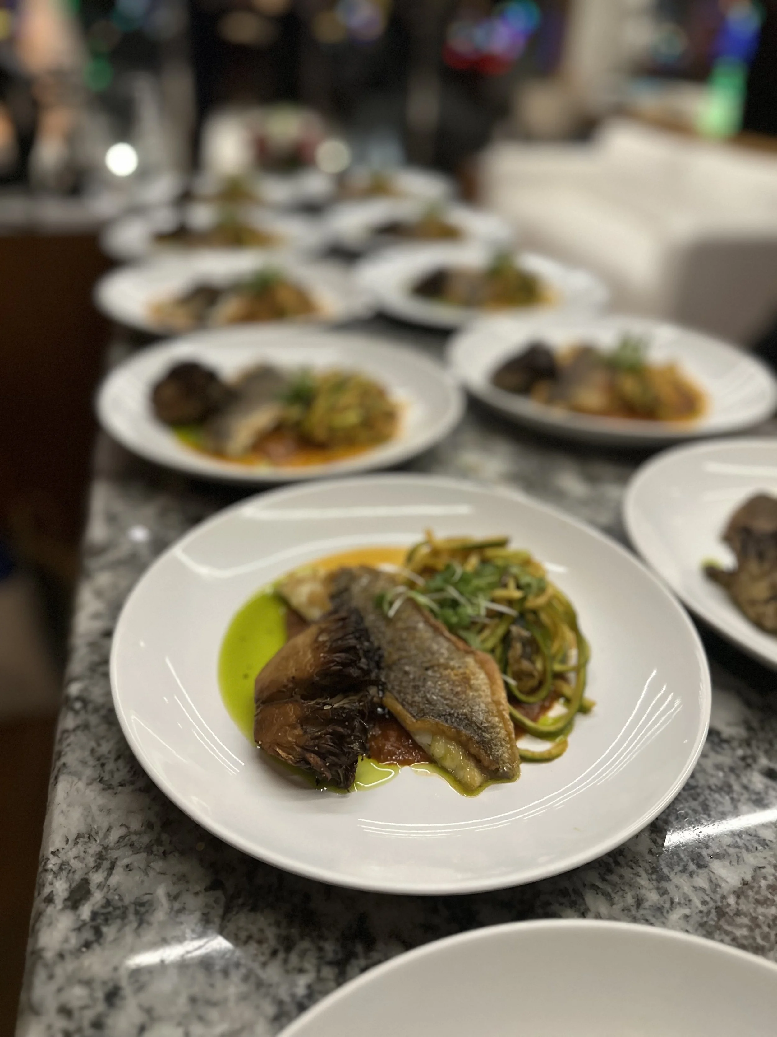 Close-up of a plated meal featuring a fish fillet with herbs and oil, served with a side of vegetables, on a white plate with a speckled gray countertop underneath.