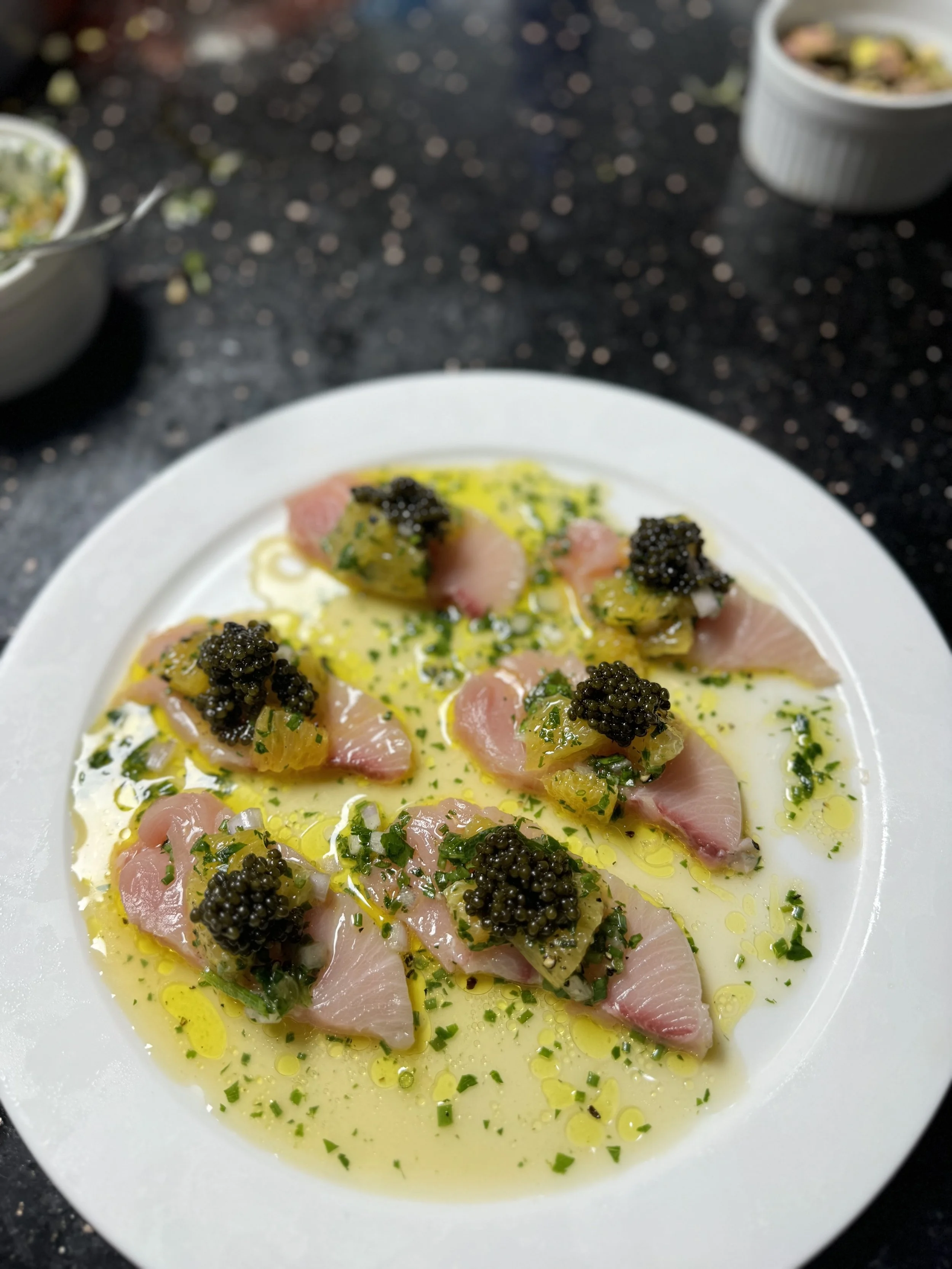 Close-up of a white plate with sliced raw fish, topped with black caviar, chopped green herbs, and drizzled with olive oil, on a dark speckled countertop.