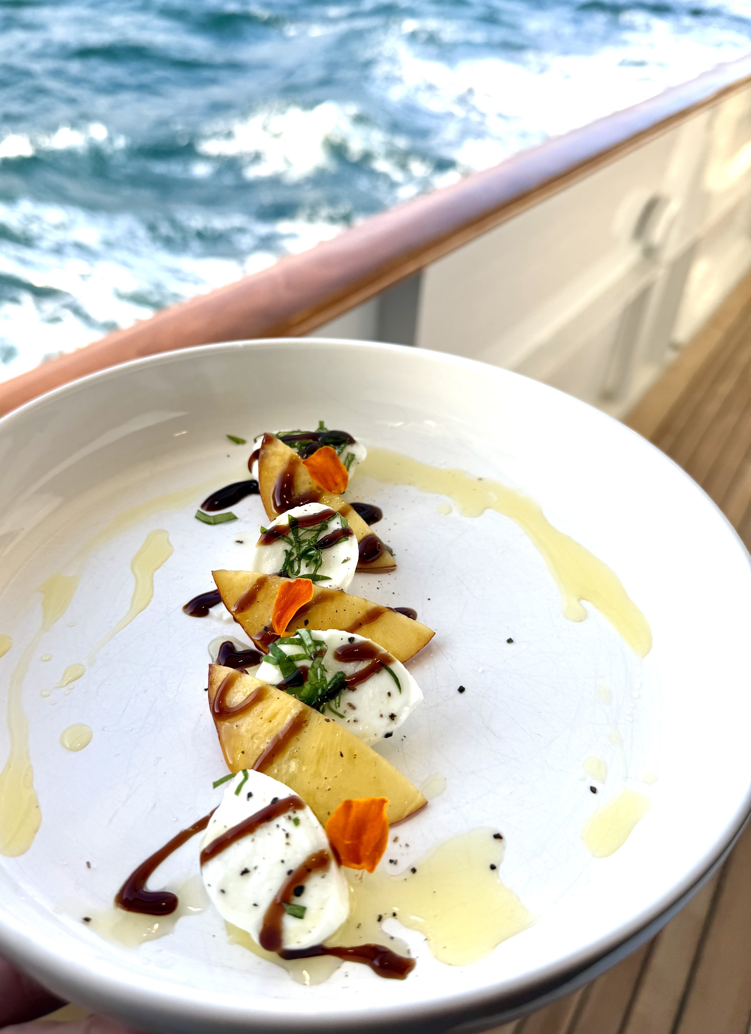 A white plate holding slices of mozzarella cheese garnished with chopped herbs, orange flower petal, and drizzled with balsamic vinegar and olive oil, with ocean waves in the background.