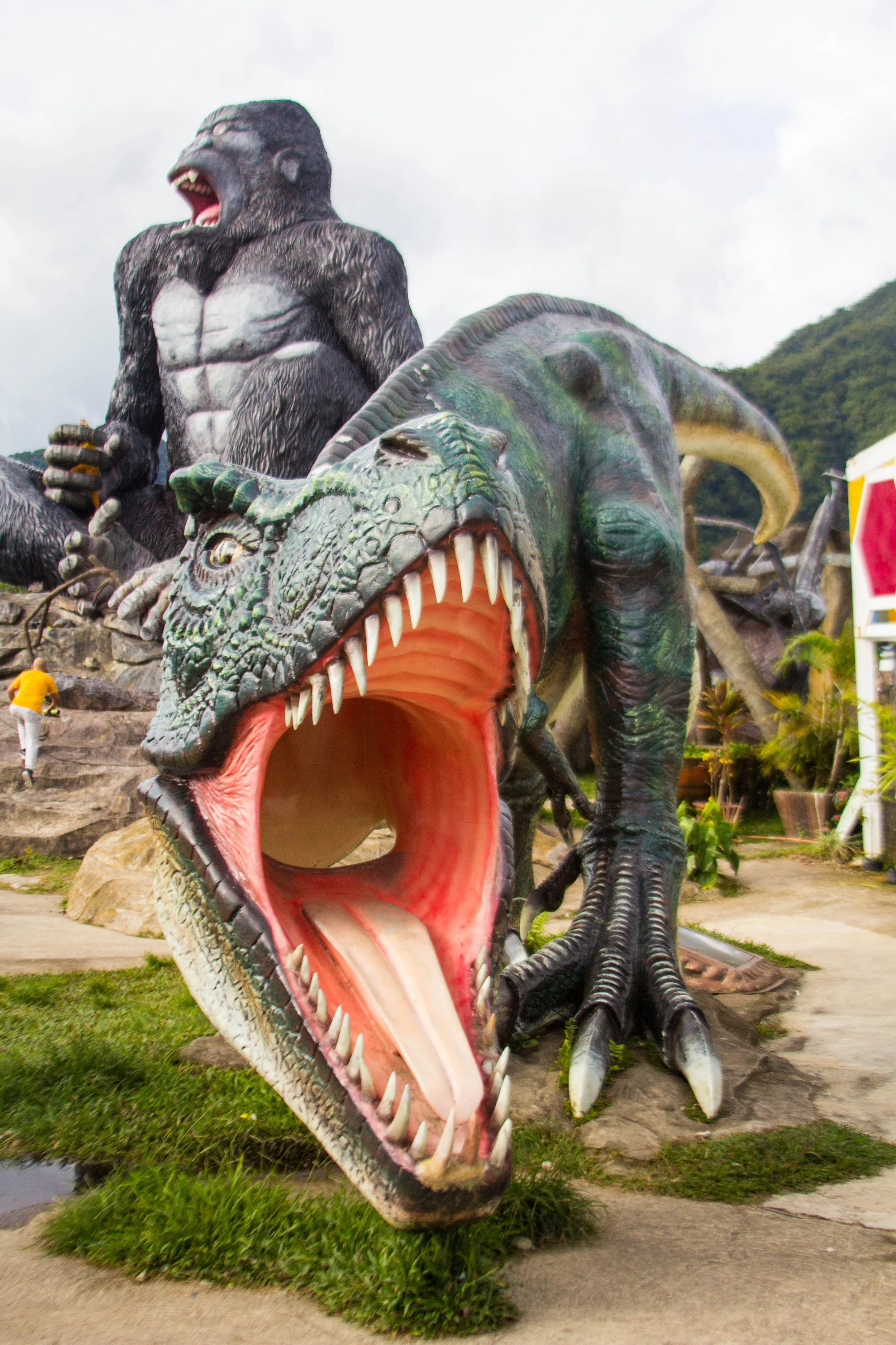 Large colorful dinosaur sculptures with an open mouth and sharp teeth, a giant gorilla figure on top, and a dinosaur skull sculpture in the foreground outdoors near mountains and greenery.