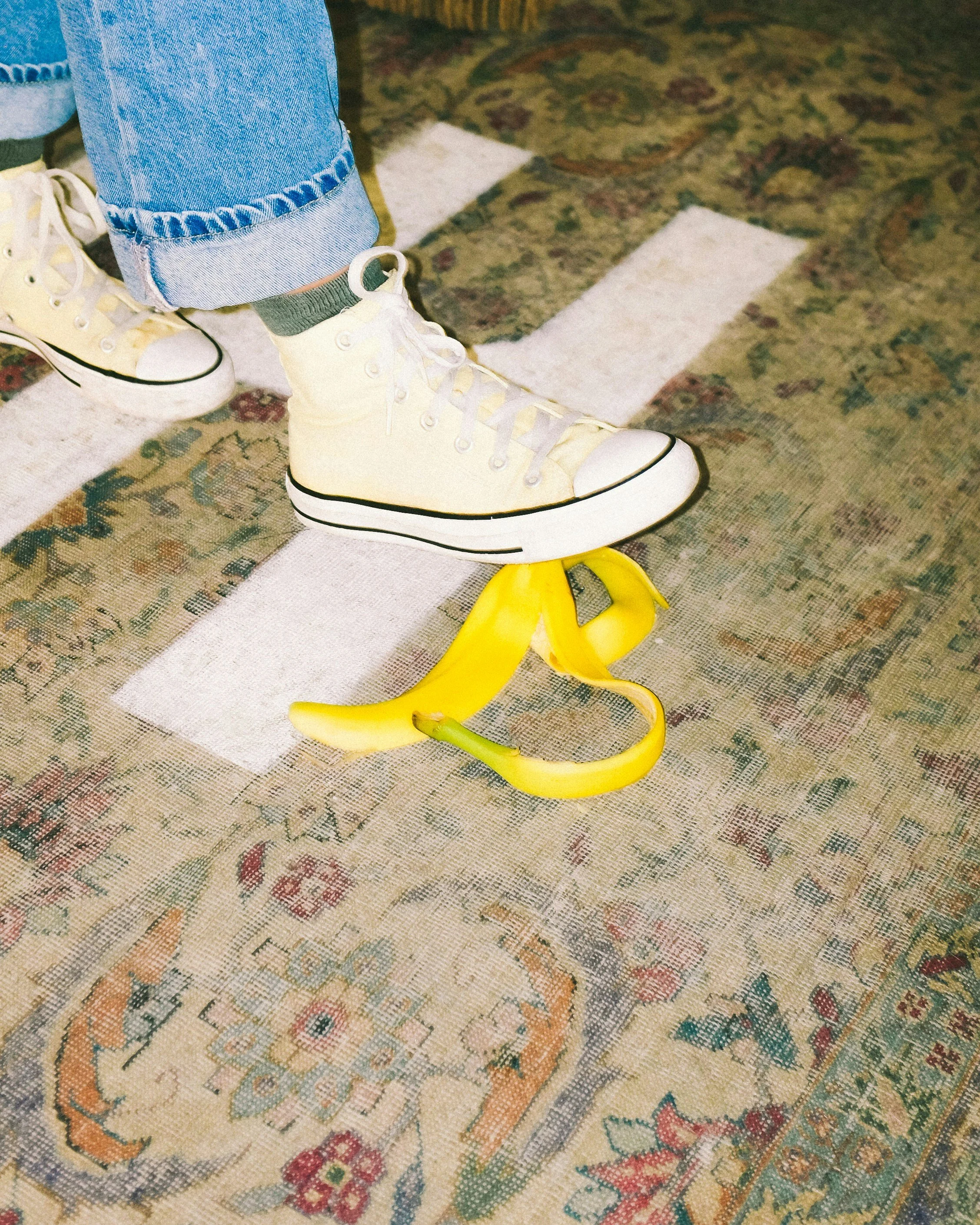 Close-up photo of a person's foot wearing a white high-top sneaker with yellow laces, standing on a yellow banana-shaped object on a patterned vintage-style carpet.