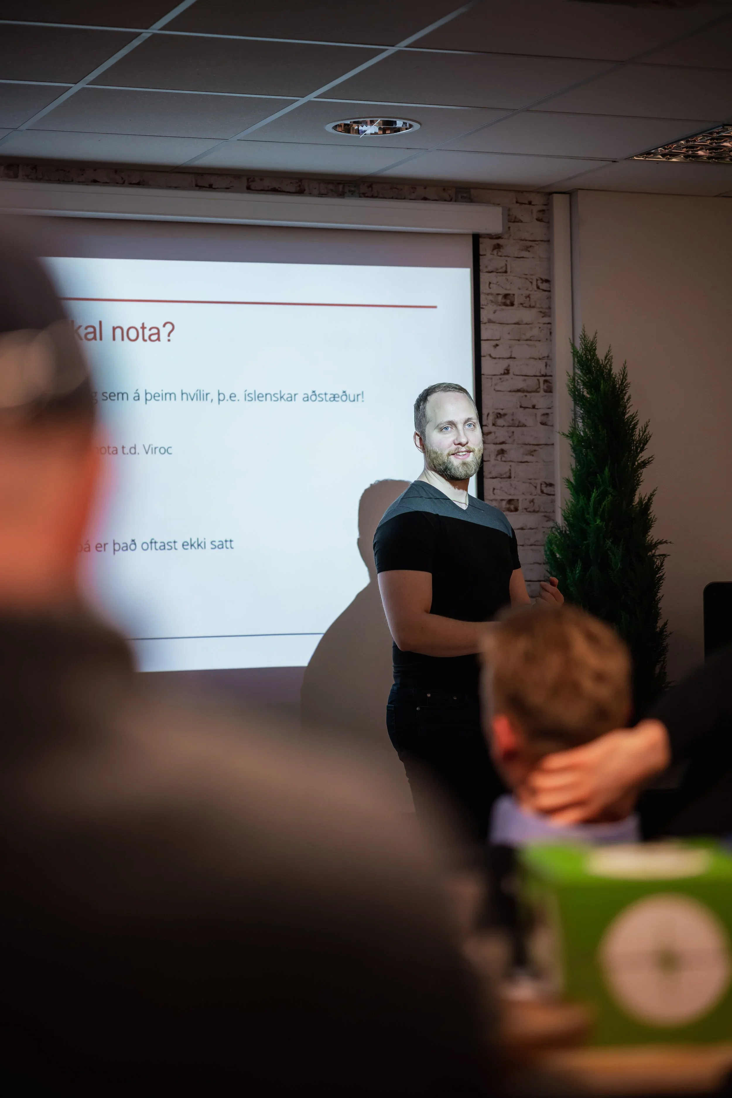 A man giving a presentation in a room decorated with a small Christmas tree. The presentation slide behind him has text in Icelandic.