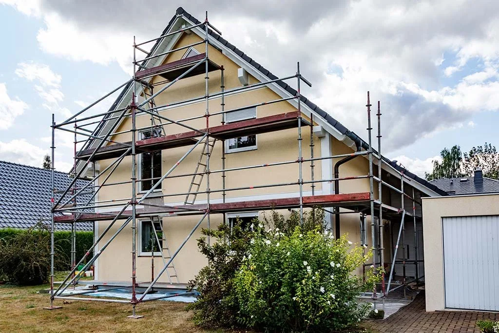 A two-story house under construction with scaffolding surrounding it, a landscaped front yard, and a garage on the right. The house has a steep roof and beige exterior walls.