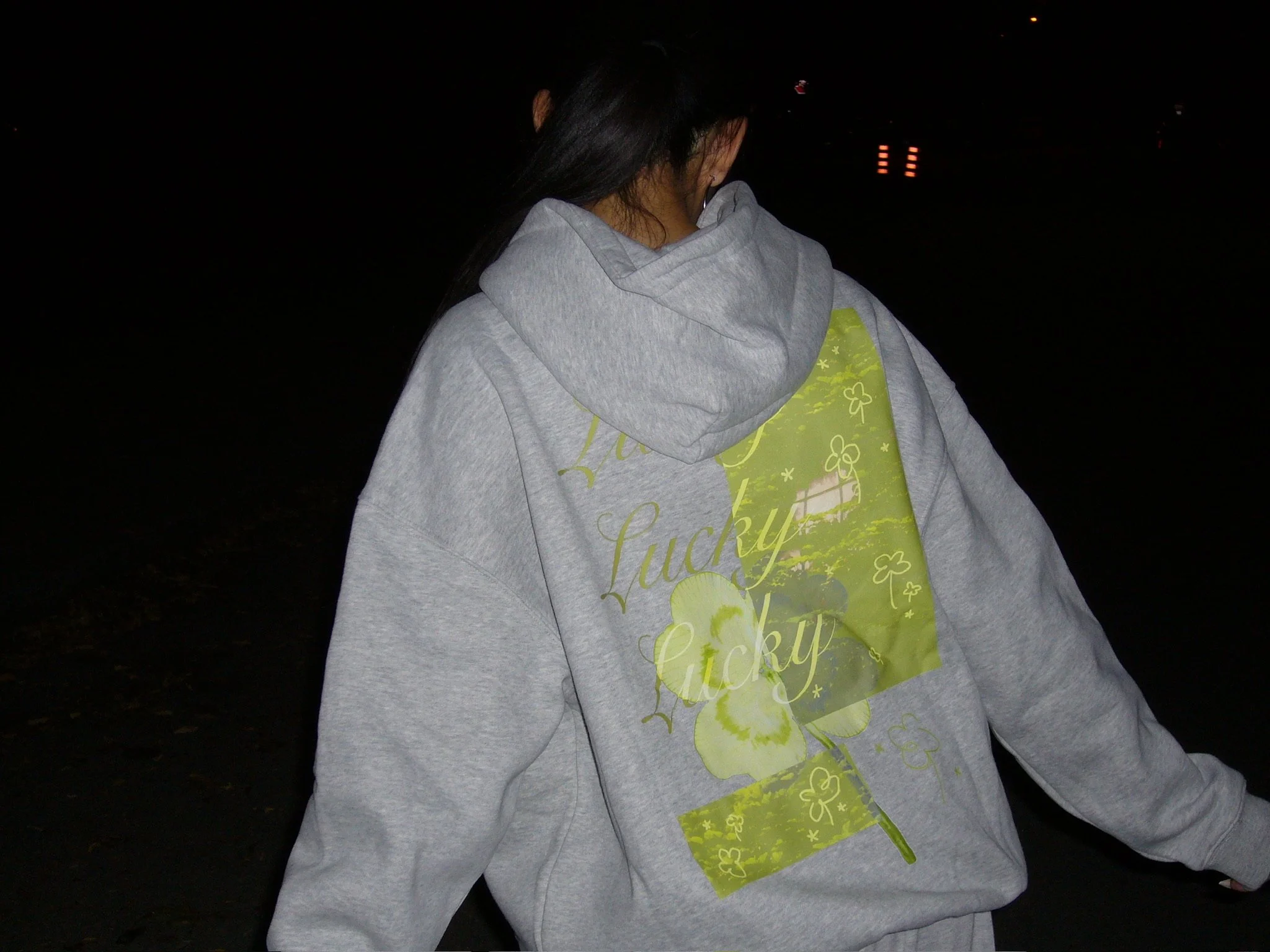 Person wearing a light gray hoodie with a green graphic design, standing outdoors at night.