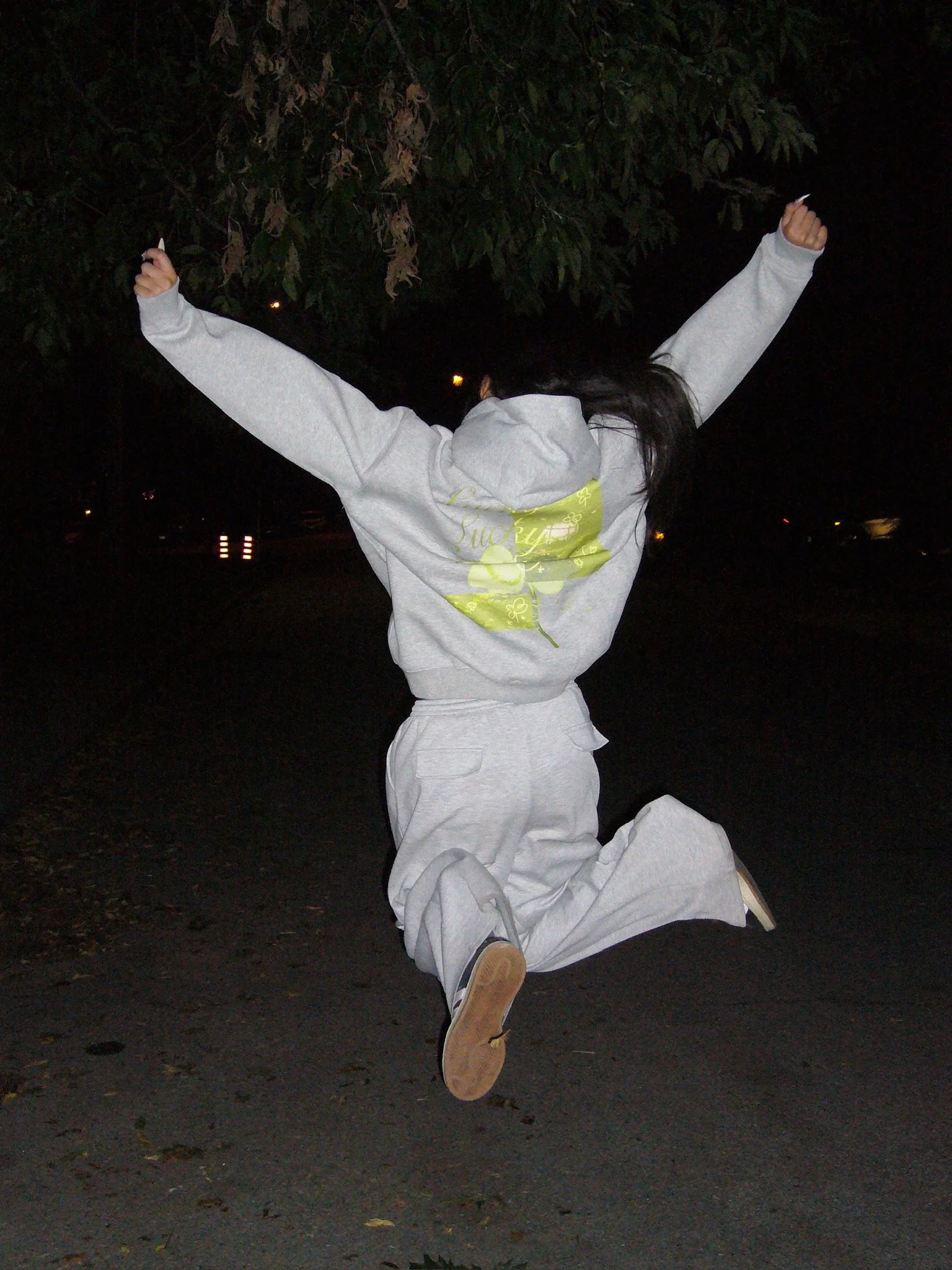 Person dressed in gray hoodie and sweatpants crouching and jumping with arms raised at night on outdoor pavement, with dark trees and faint lights in the background.