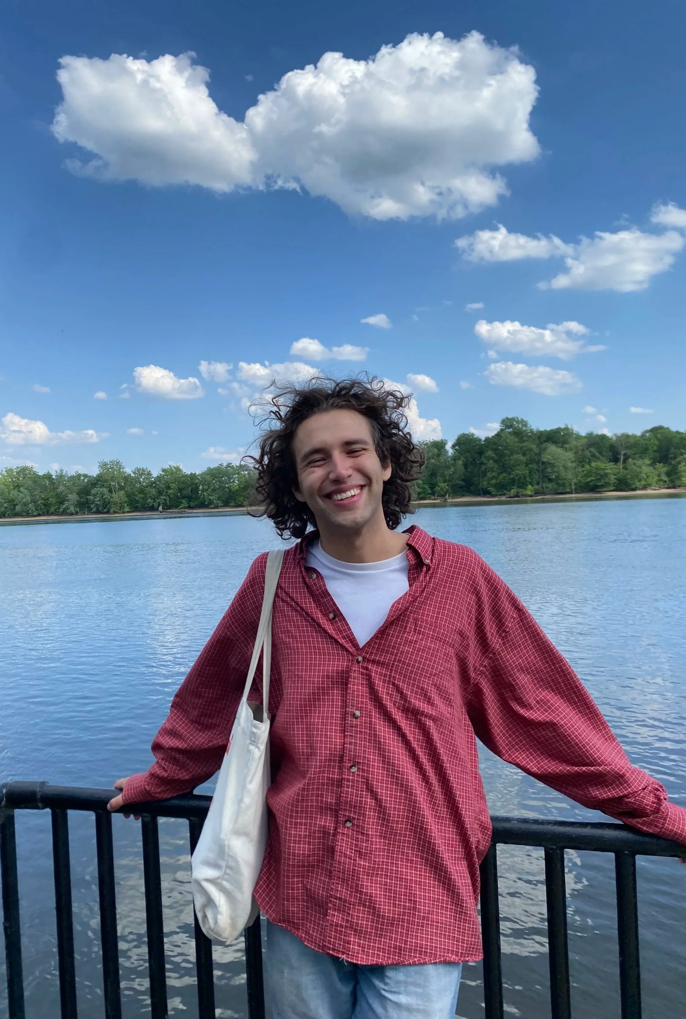 A young man with curly hair wearing a red checkered shirt and blue jeans, standing by a water body with trees in the background, smiling at the camera under a blue sky with white clouds.