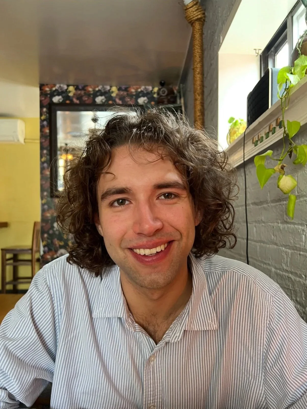 A young man with curly brown hair, smiling and wearing a white striped shirt, sitting in a cafe with a colorful floral wallpaper and potted plants.