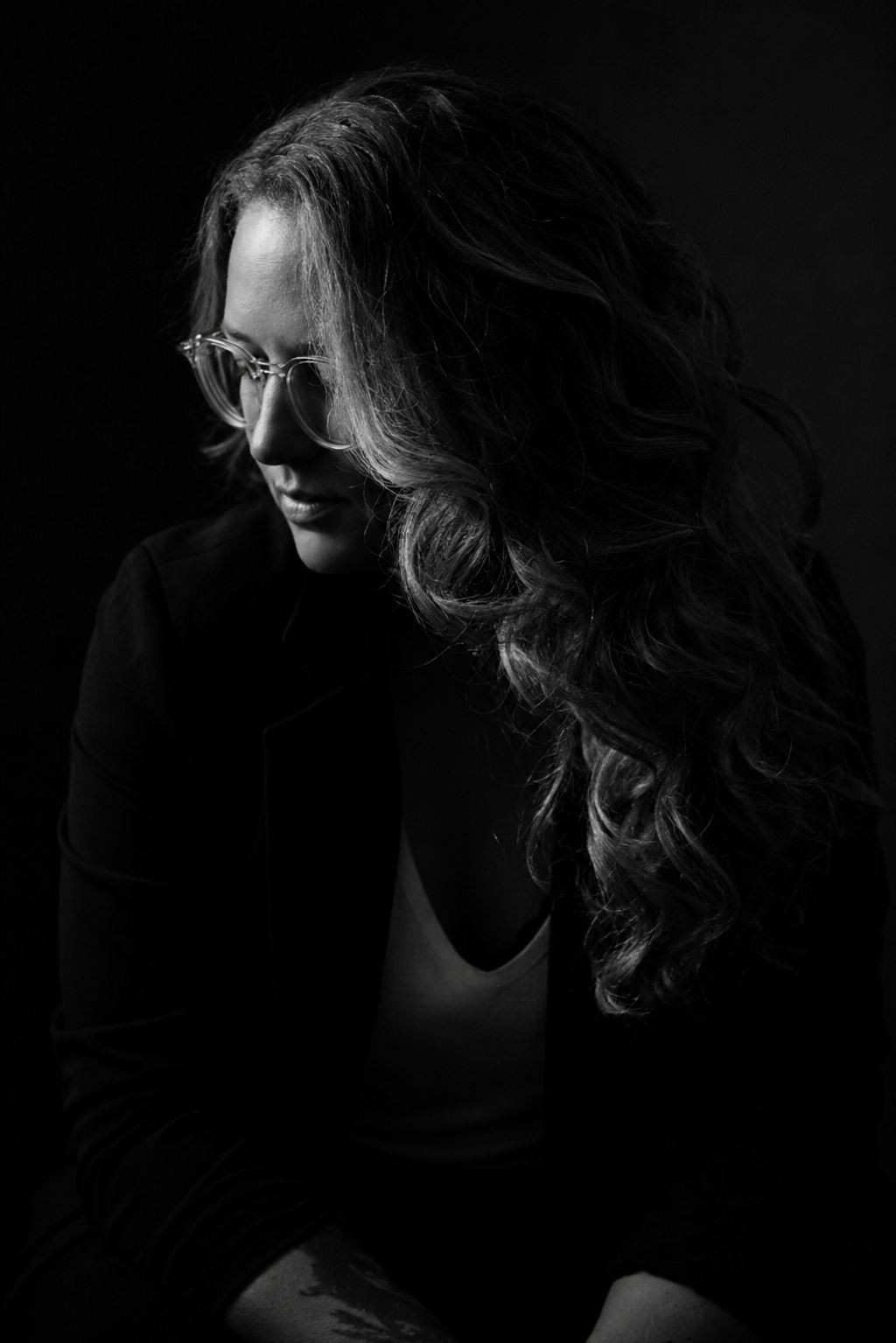 Black and white portrait of a woman with long, curly hair, wearing glasses, looking down, against a dark background.