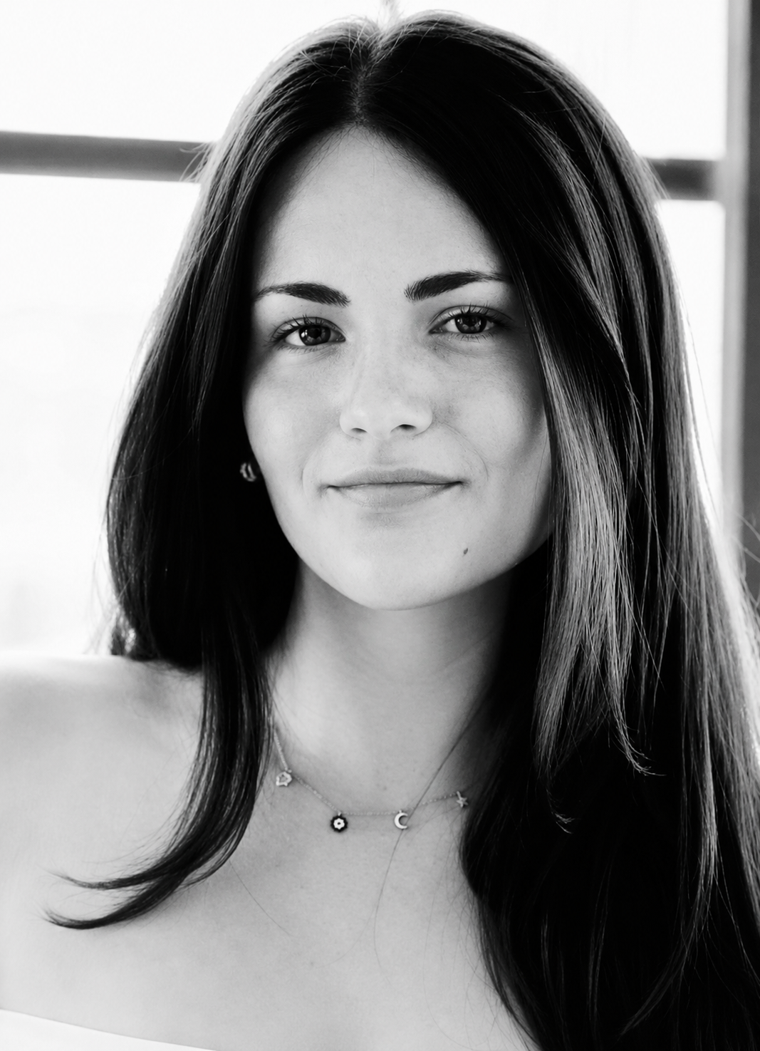 Black and white portrait of a young woman with long dark hair, wearing multiple necklaces, smiling softly.