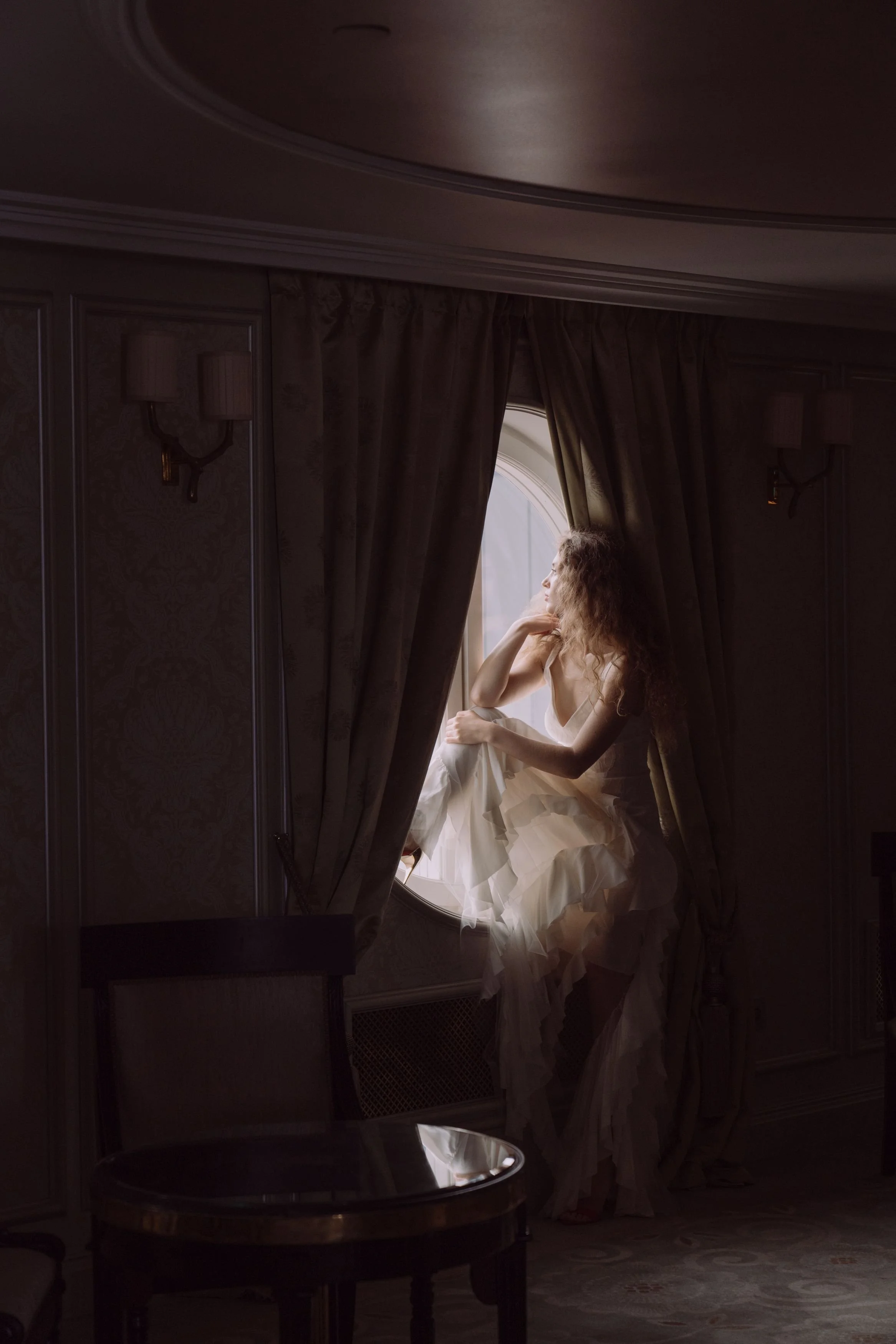 A woman with curly hair in a white dress sitting by a round window with her hand on her chin, gazing outside, in a dimly lit room with dark curtains and ornate wallpaper.