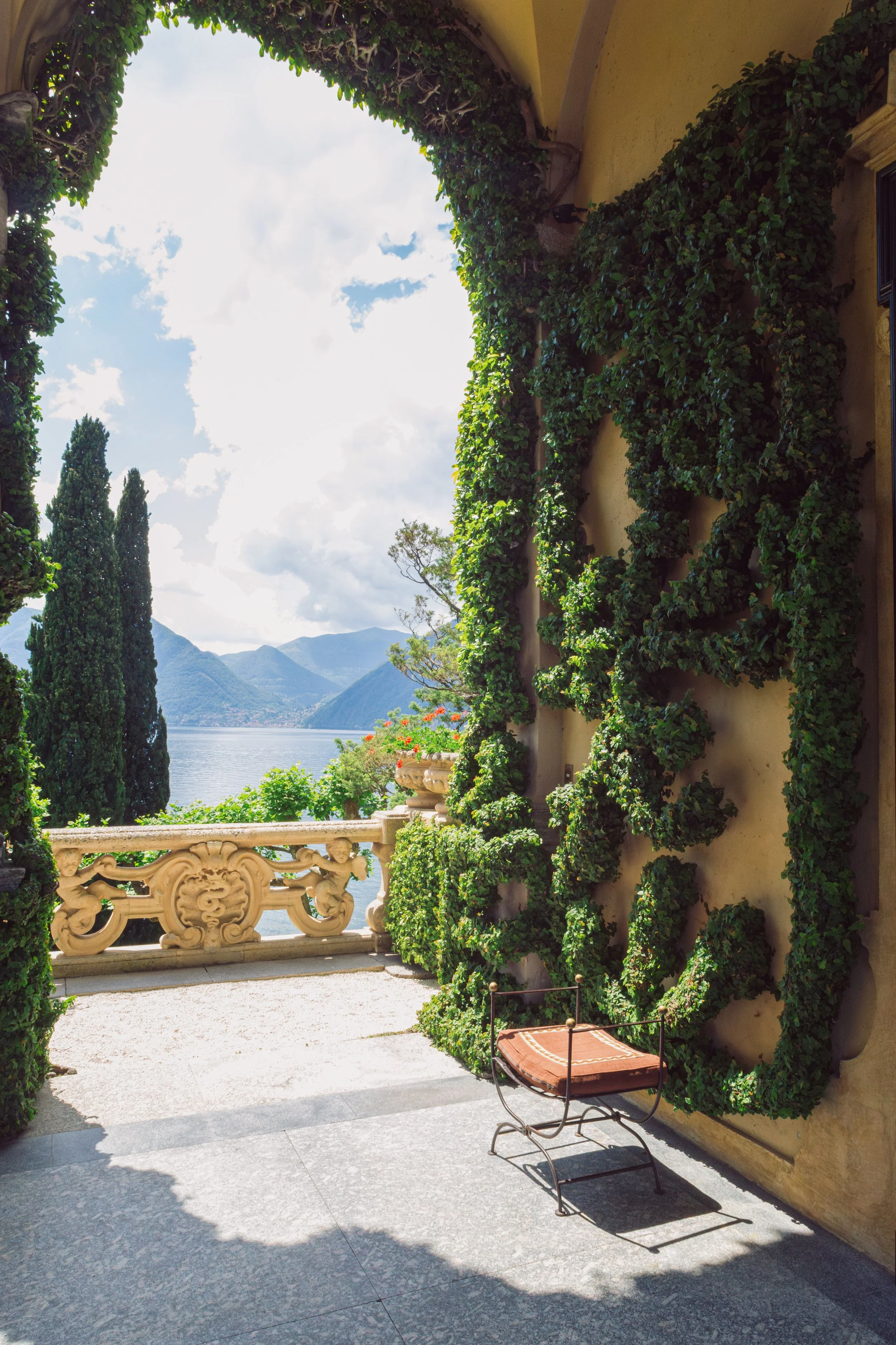 A scenic view of a terrace with stone flooring, partially shaded, overlooking a lake with mountains in the distance. The terrace has decorative stone railings and lush greenery, including ivy climbing a wall, and tall trees in the background. There is a vintage-style chair with a cushioned seat in the foreground.