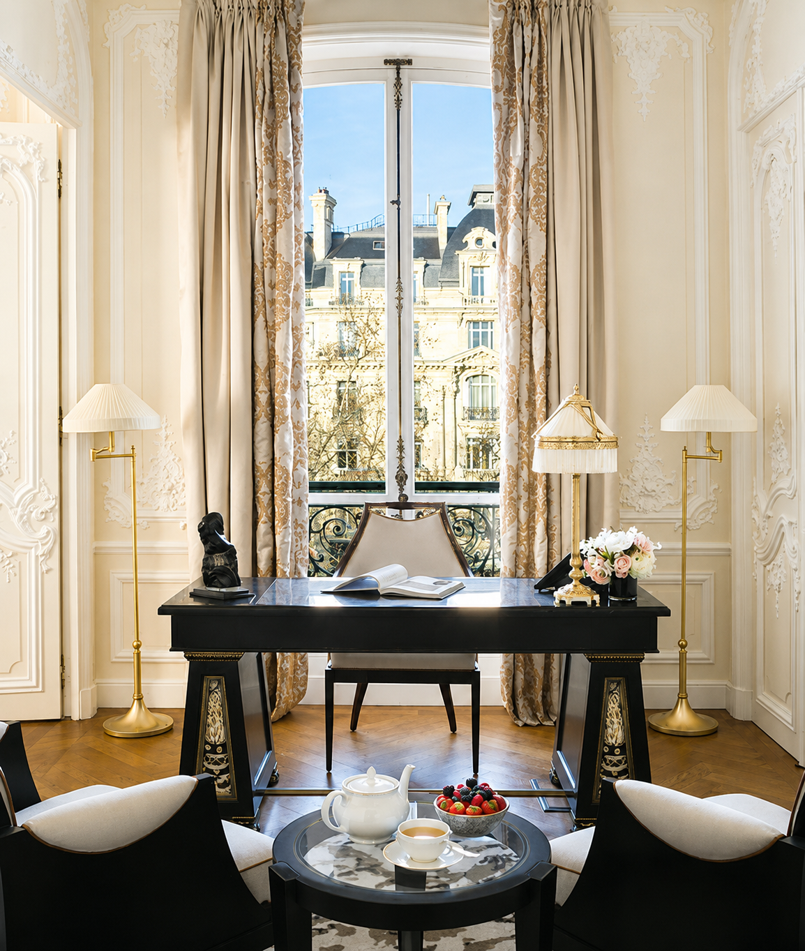 Elegant hotel room with a large window, ornate drapes, a black desk with a book and decorative objects, cream-colored walls with decorative molding, two lamps, a tea set and strawberries on a round table, and a view of historic buildings outside.