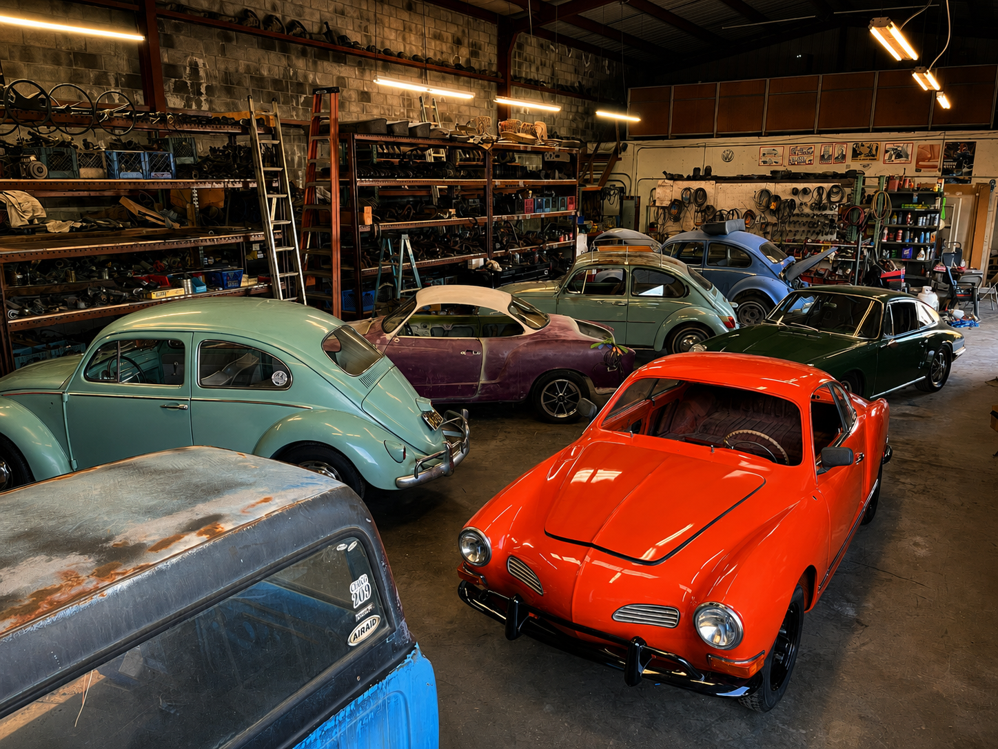 Interior of a garage filled with vintage Volkswagen Beetle cars in various colors, including orange, green, purple, and blue, with shelves and tools in the background.