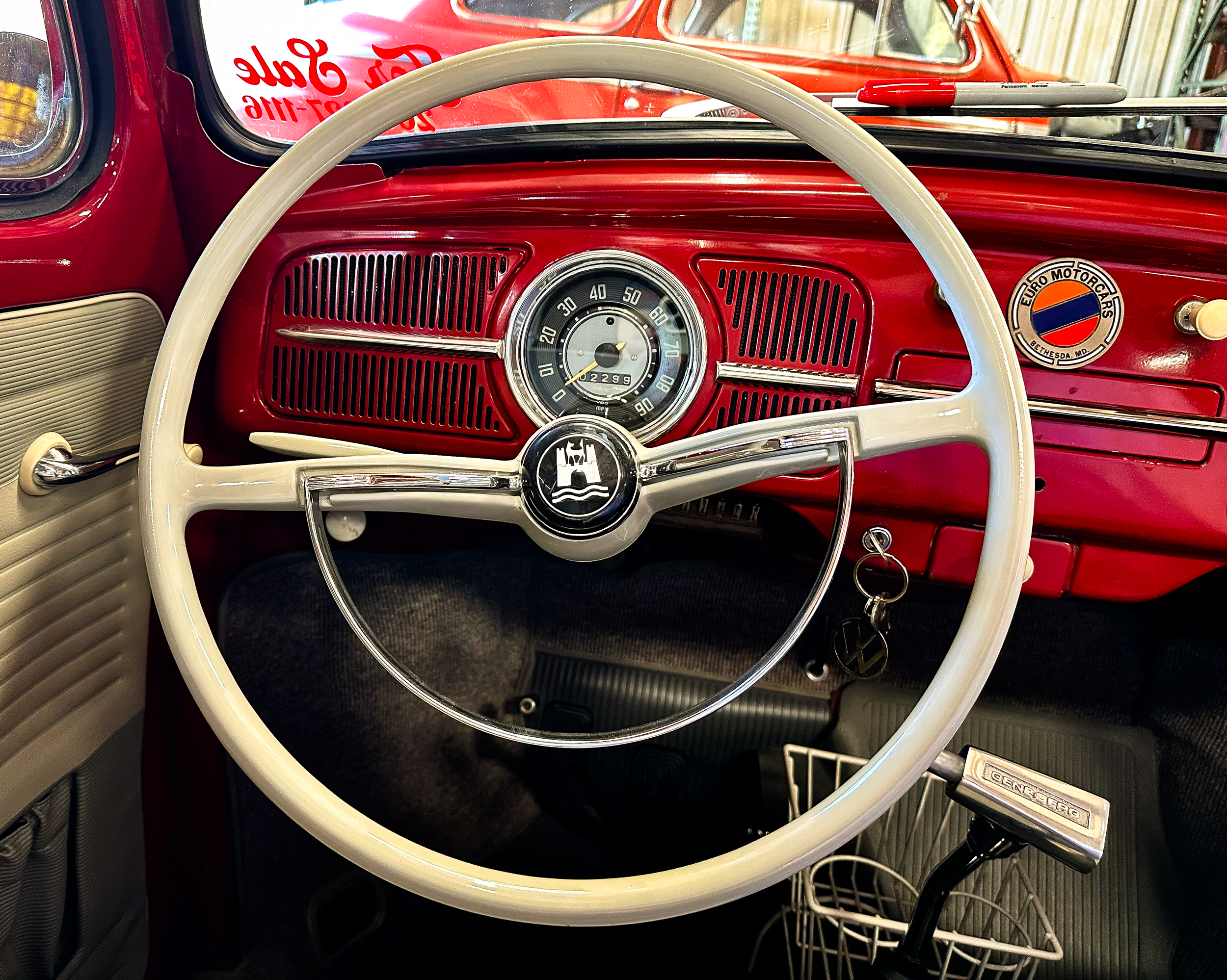 The image shows the interior of a vintage red car, focusing on the dashboard and steering wheel. The dashboard has a circular speedometer in the center, and a Euro Motorcars badge on the right. A red and gray permanent marker is resting on the dashbo
