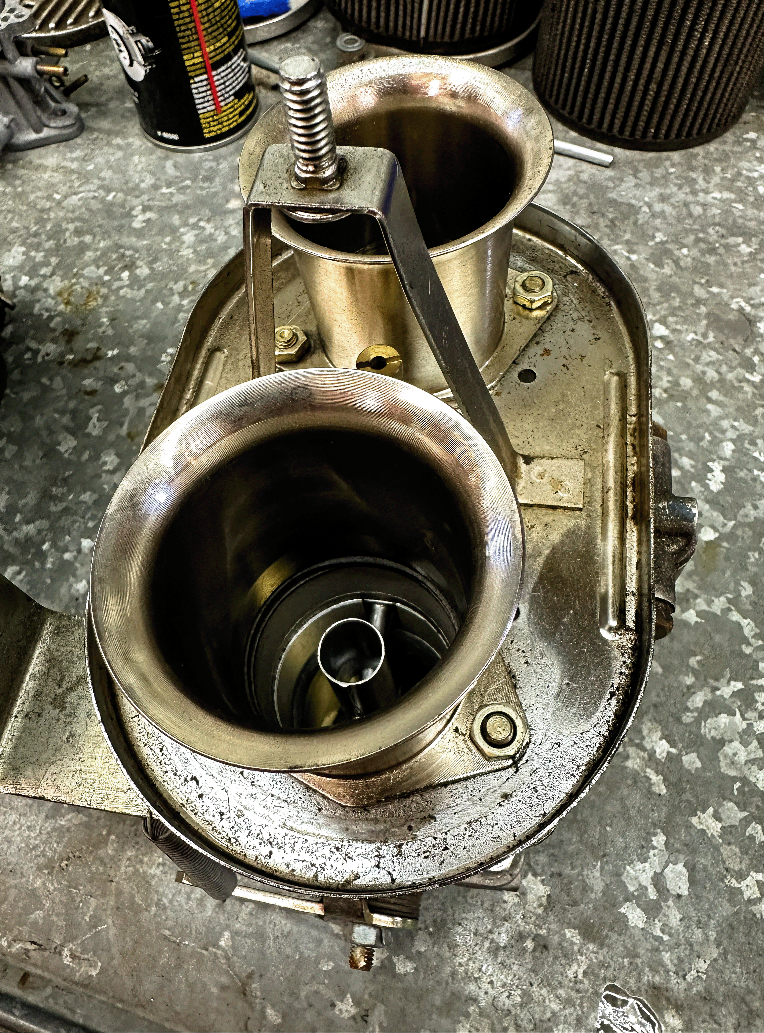 Close-up view of a mechanical part, likely a turbocharger or similar engine component, with metallic circular openings and a spring mechanism, surrounded by a work surface with tools and supplies.