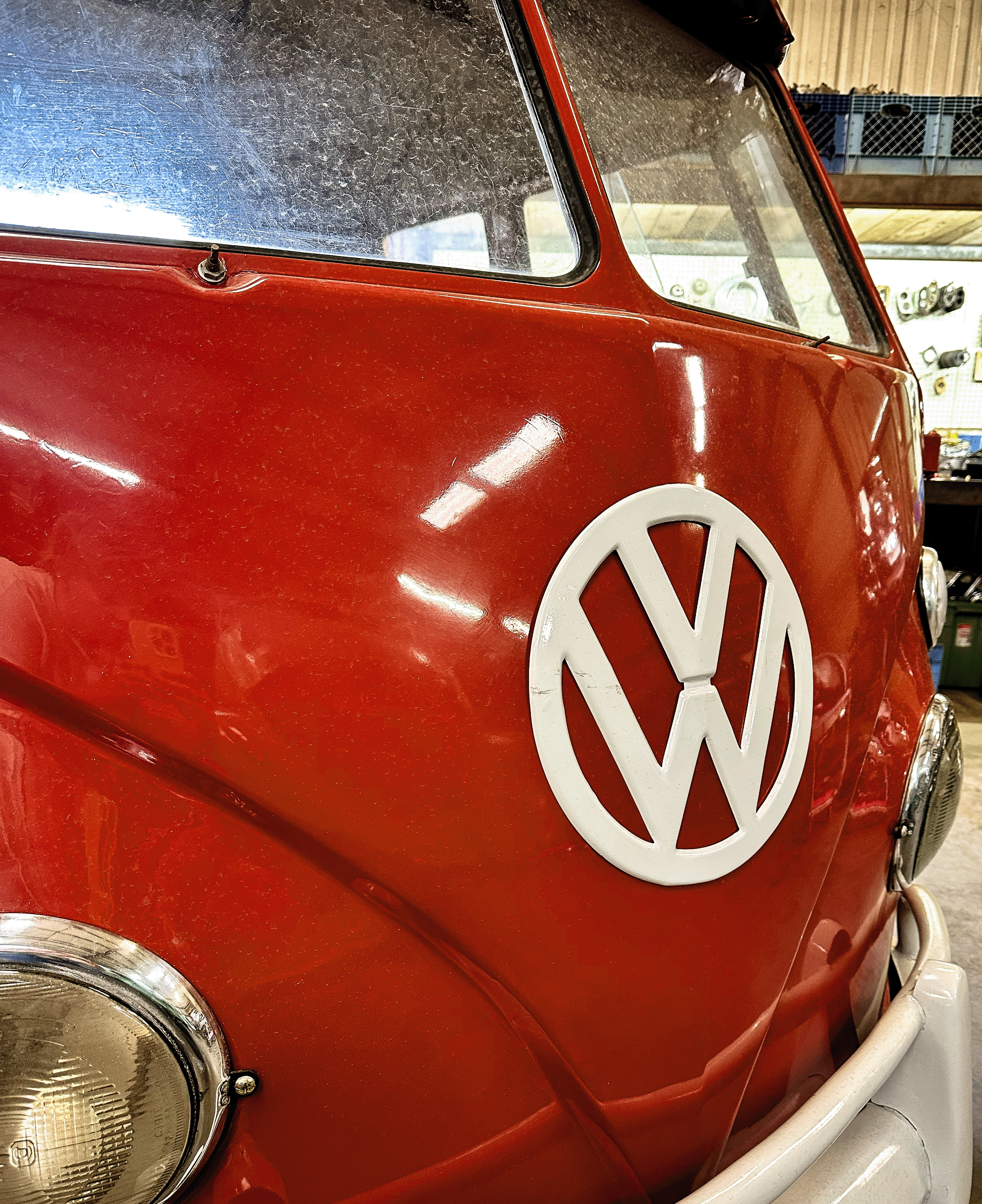 Close-up of a red vintage Volkswagen van with a large VW emblem on the front, parked in a garage with tools and supplies on the wall behind.