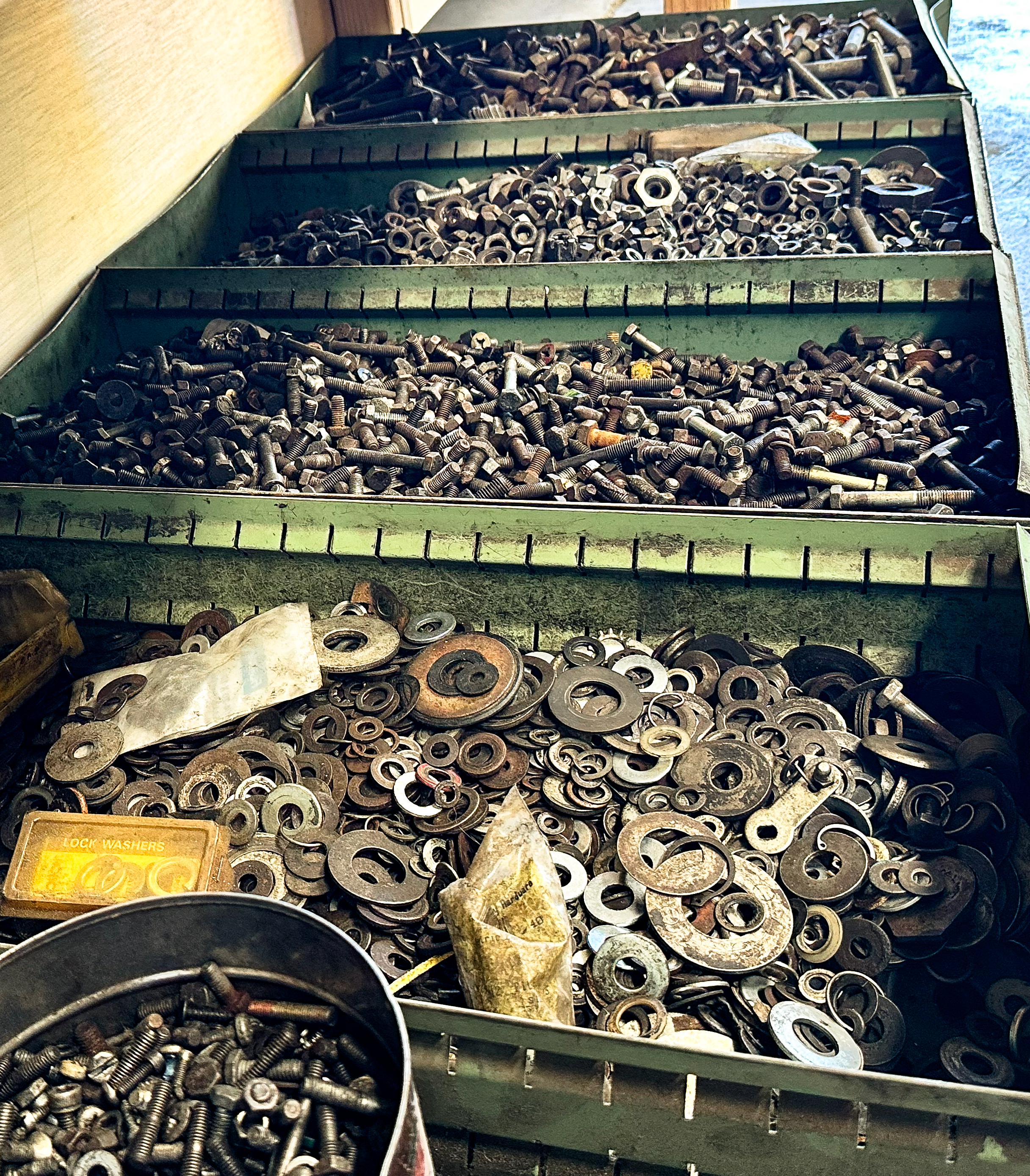 Three green metal trays filled with various metal bolts, nuts, and washers. The bottom tray has a mixture of rusted and shiny metal washers and bolts, some in a black container and others loose. The trays are on a shelf or counter.