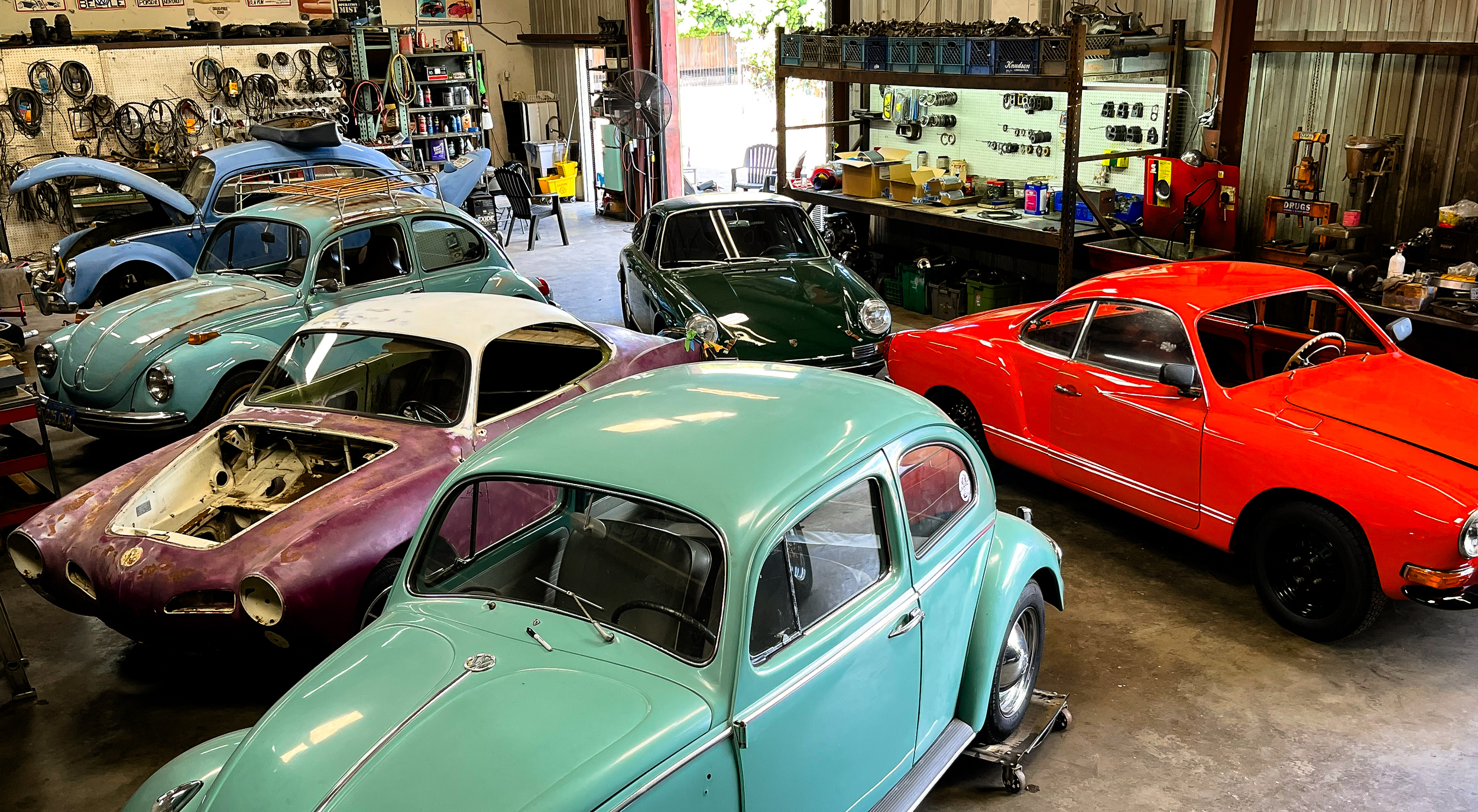 Several vintage Volkswagen Beetle cars in various colors, including green, black, blue, and red, are inside a workshop. Some cars are partially disassembled, and tools and parts are visible on shelves and workbenches.